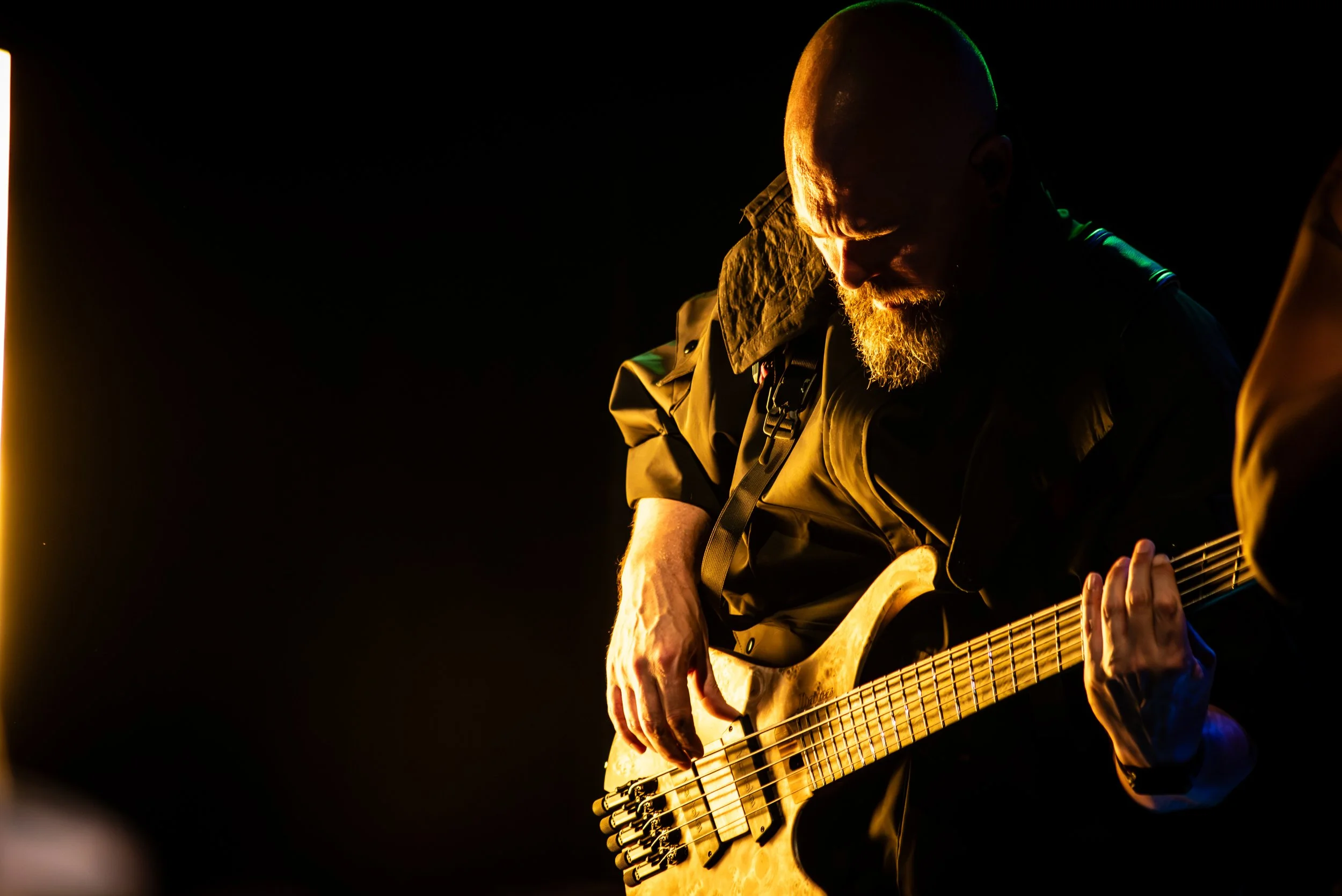 A man with a beard, wearing glasses and a dark jacket, playing an acoustic guitar under warm stage lighting.