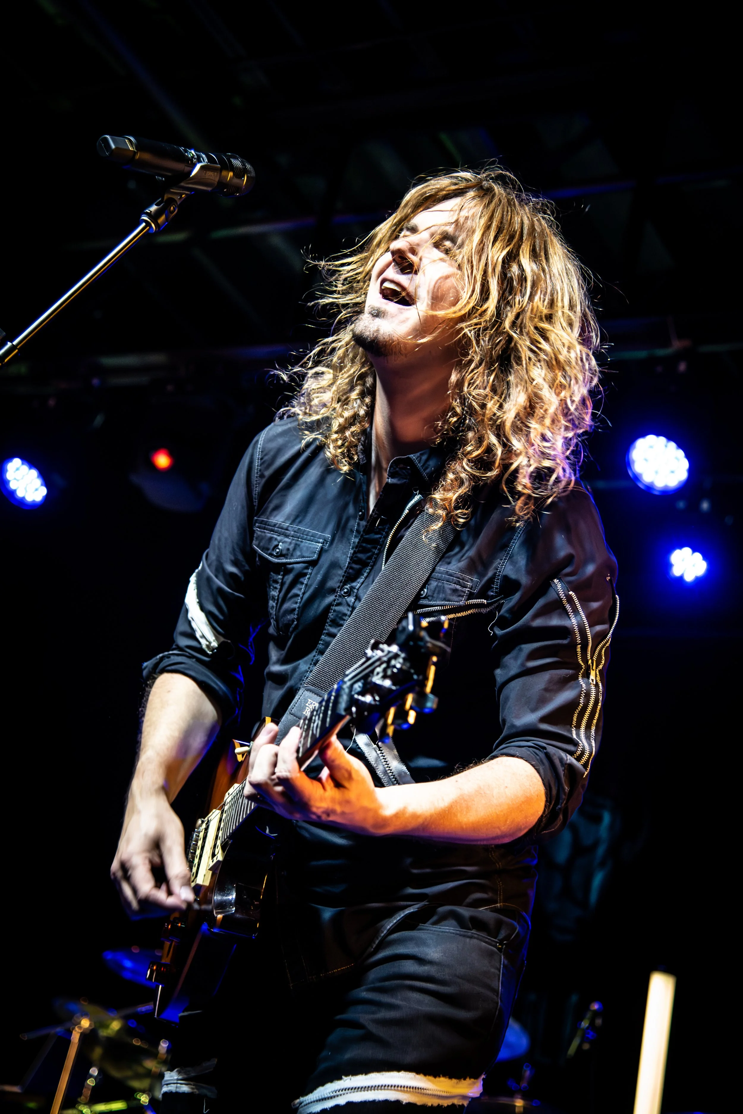 A male musician with long curly hair passionately singing into a microphone while playing an electric guitar on a stage with colorful lights.