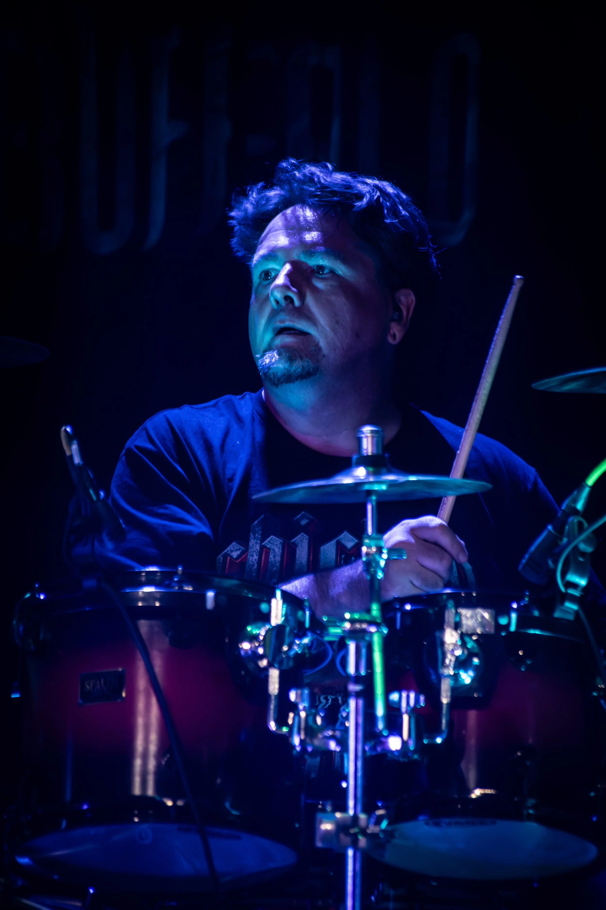 A man with wavy hair and a goatee playing a drum kit on stage, illuminated by blue and purple stage lights.