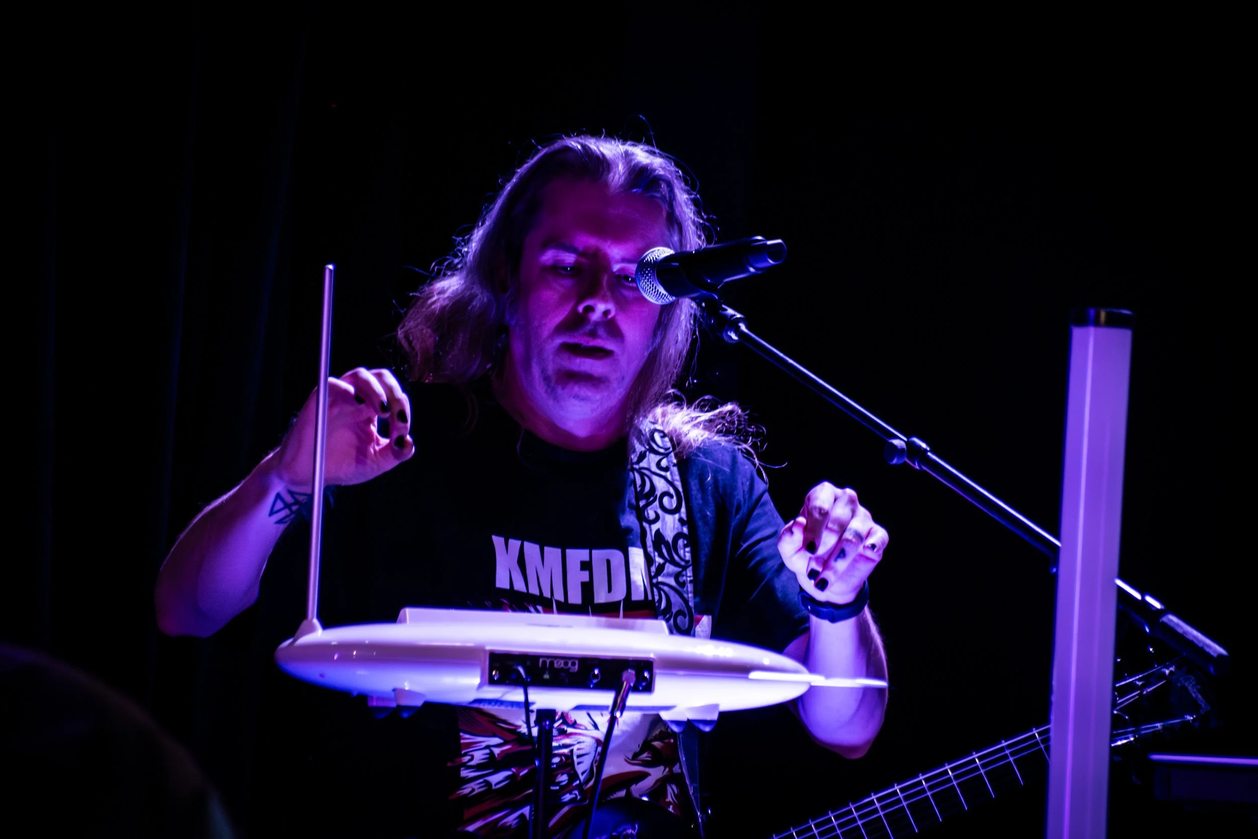 Person with long hair performing with electronic musical equipment on a dark stage, illuminated by purple lighting.