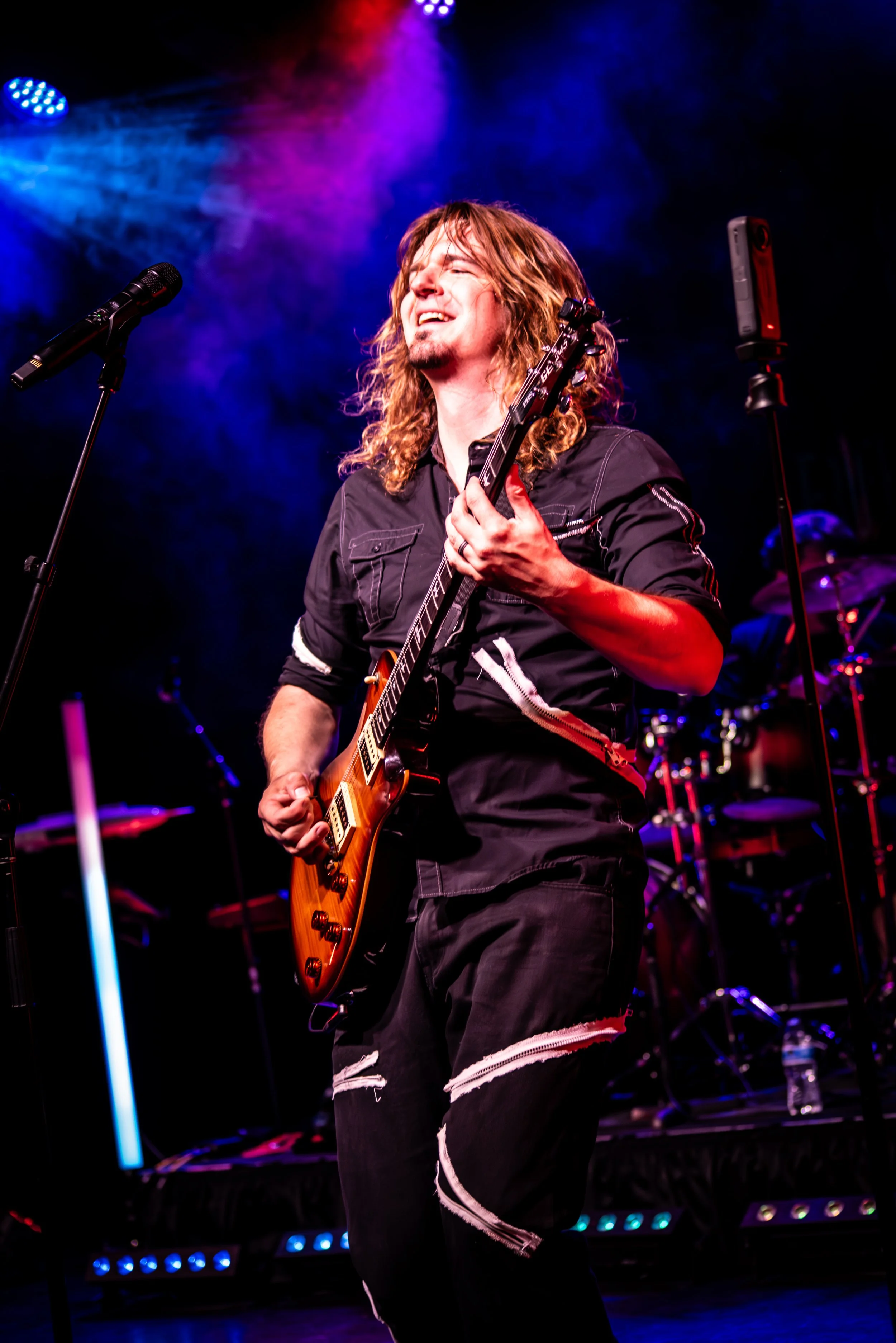 A man with long curly hair and a goatee playing an electric guitar on stage, smiling with eyes closed, under colorful stage lights.