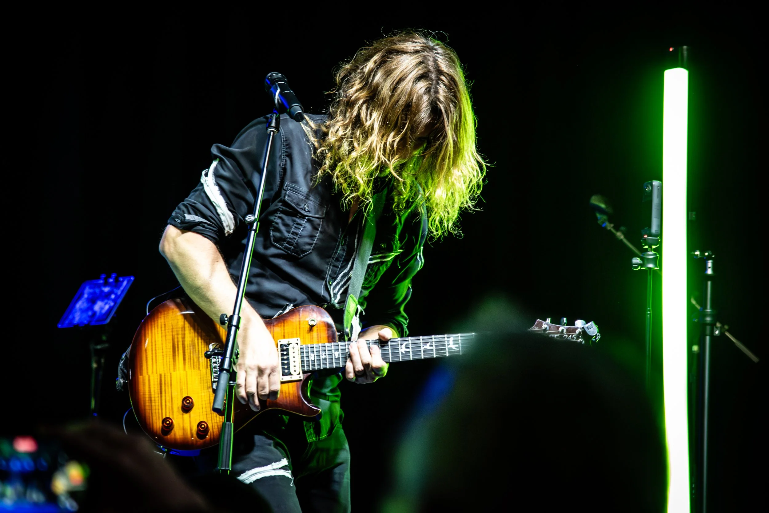 Musician with curly hair playing an electric guitar on stage, illuminated by green and white stage lights, with equipment visible in the background.