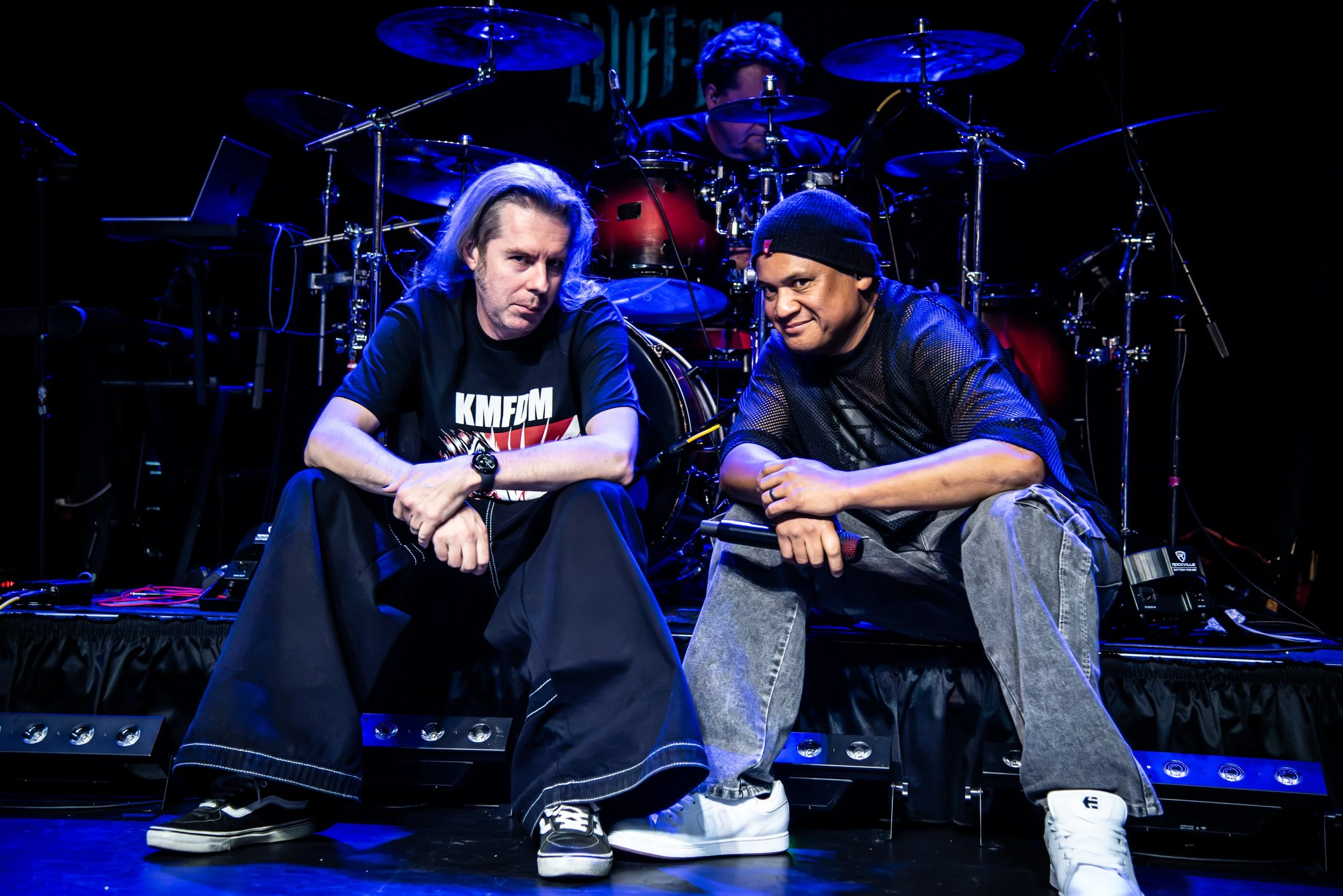 Two male performers sitting on stage with a drum set and equipment behind them, illuminated by blue stage lighting.
