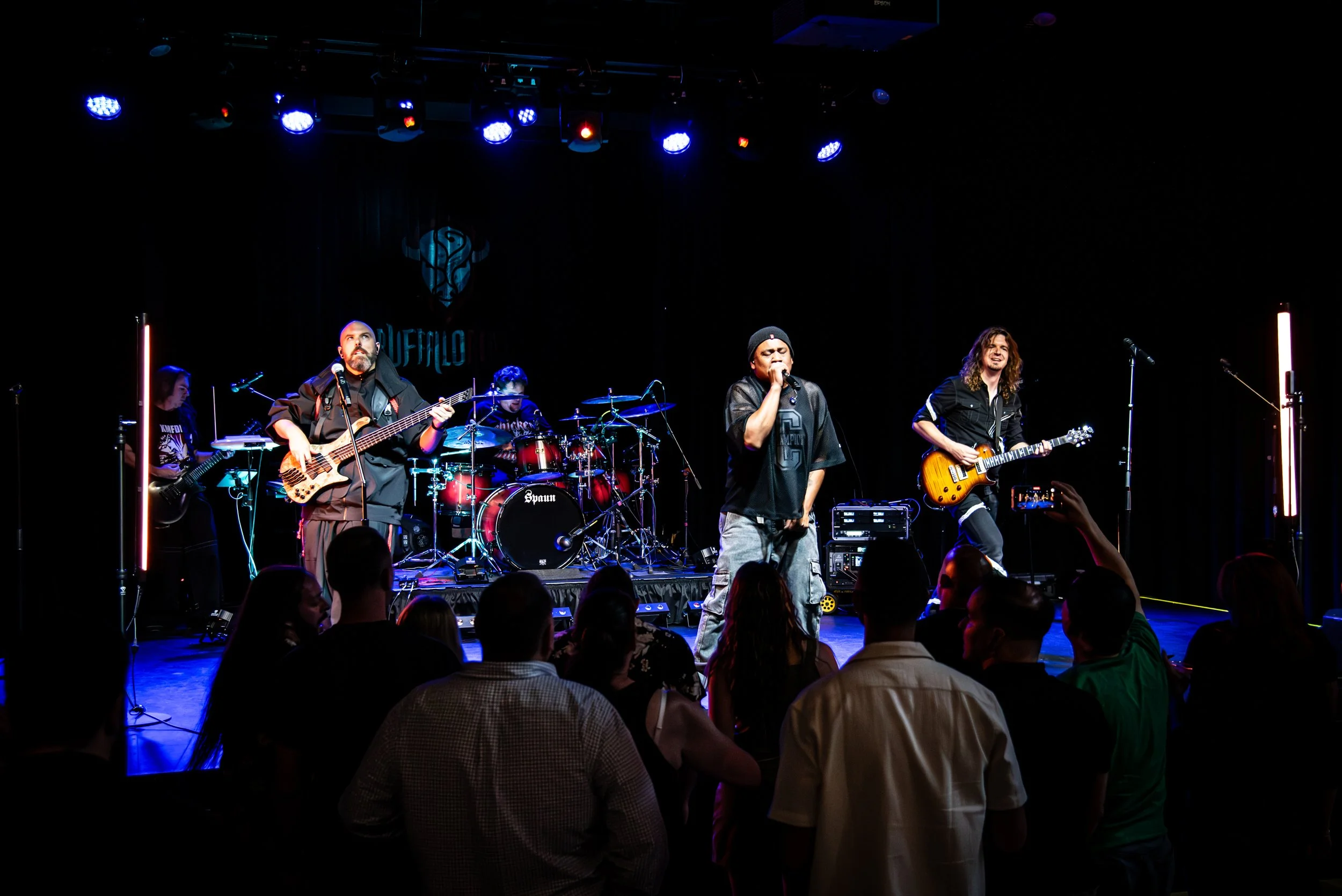 A band performing on stage with five members, with the audience in front, under blue stage lighting.