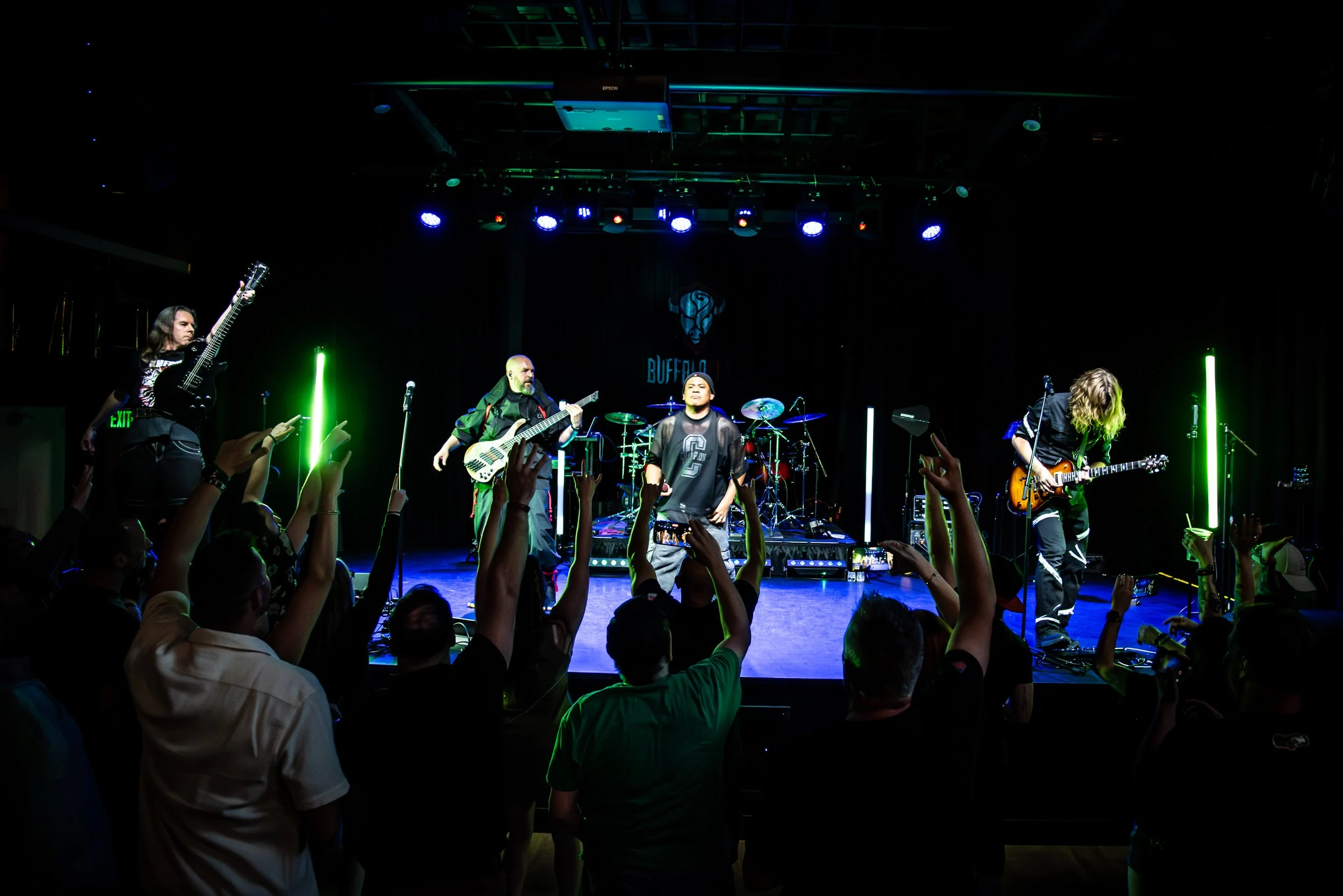 A live music performance on stage with four musicians playing guitars and drums, audience members raising hands, stage lighting in blue and green, venue sign 'BUFFALO' in the background.