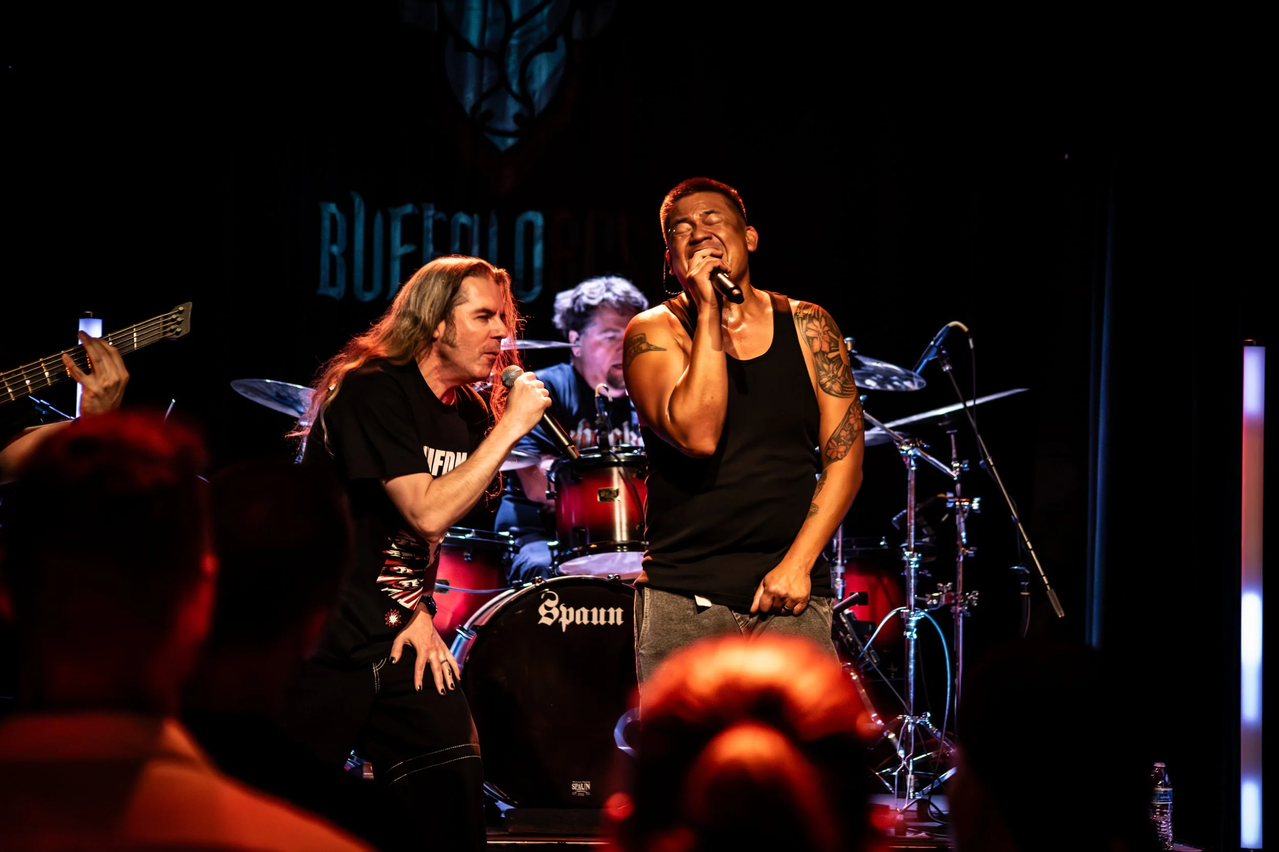 A band performs on stage, with a woman singing passionately into a microphone, wearing a black tank top with tattoos on her arm, and a man with long hair also singing with a microphone. In the background, a drummer is visible behind a drum set, and t