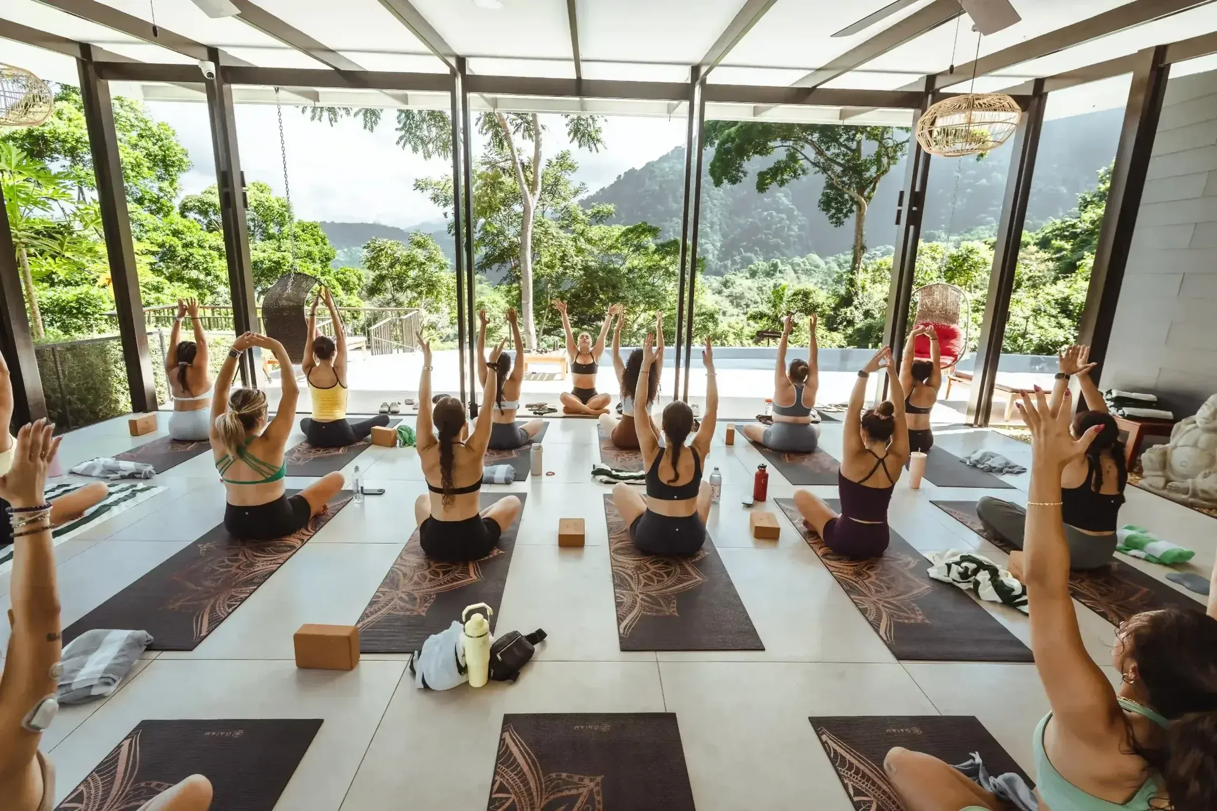 A group of women practicing yoga in a spacious, glass-enclosed studio with outdoor mountain views, seated on yoga mats with their arms raised.