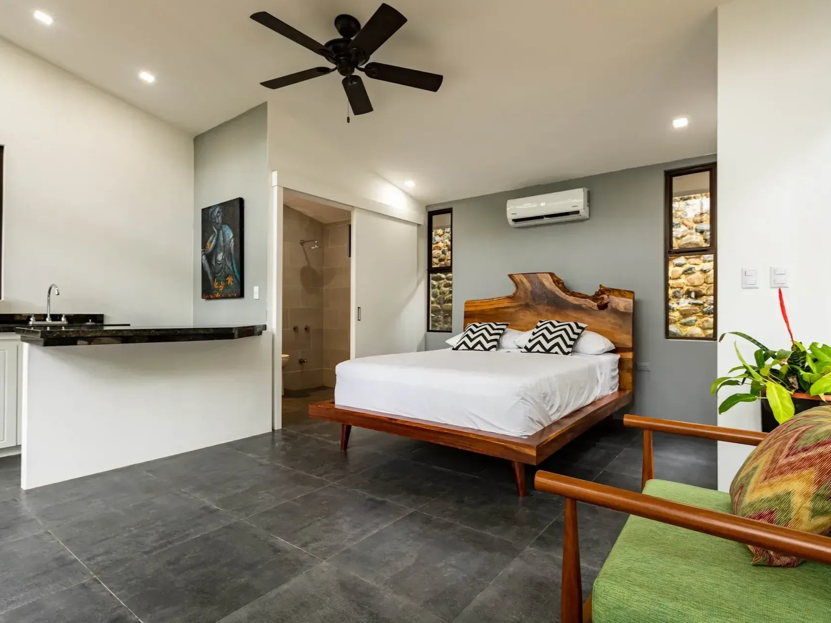 A modern bedroom with a wooden bed frame, white bedding, and decorative pillows with a black and white chevron pattern. There is an air conditioning unit above the bed, two narrow windows with decorative stone views, a ceiling fan, and a partial view of a bathroom with a shower. A green upholstered chair with a floral pillow is in the foreground, and a potted plant is on the right side.