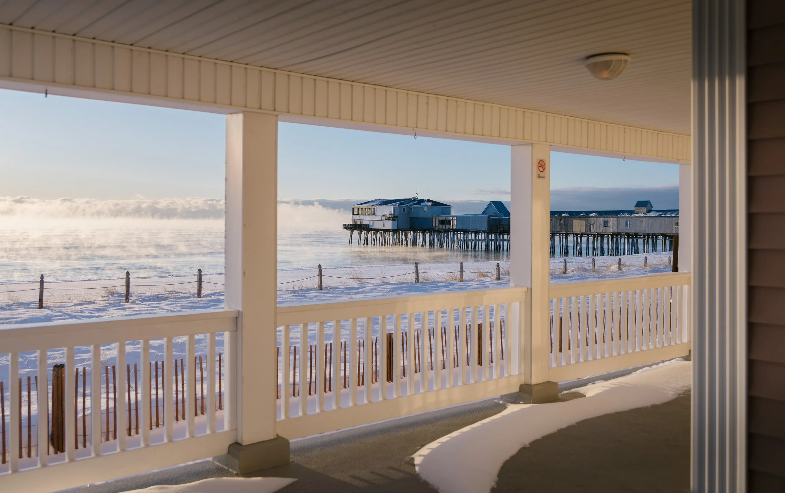 View from a covered porch overlooking a snowy beach with a pier extending into the water and a building at the end of the pier during winter.
