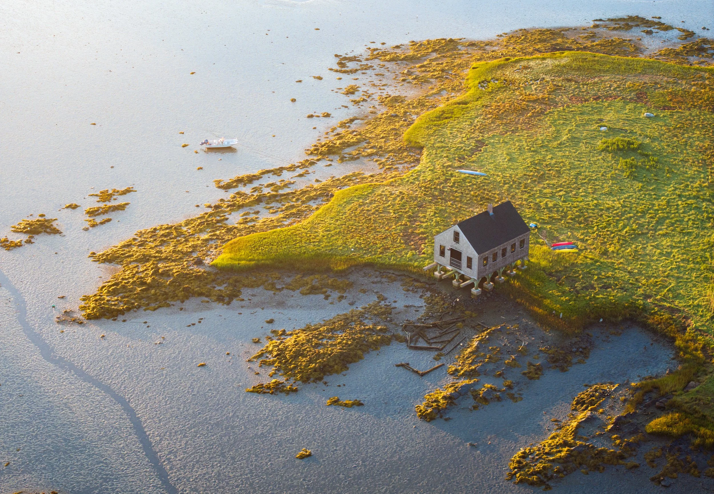 An aerial view of the fish house in Kennebunkport on stilts near a body of water with patches of grass and moss, a boat docked nearby, and other small boats on the land.