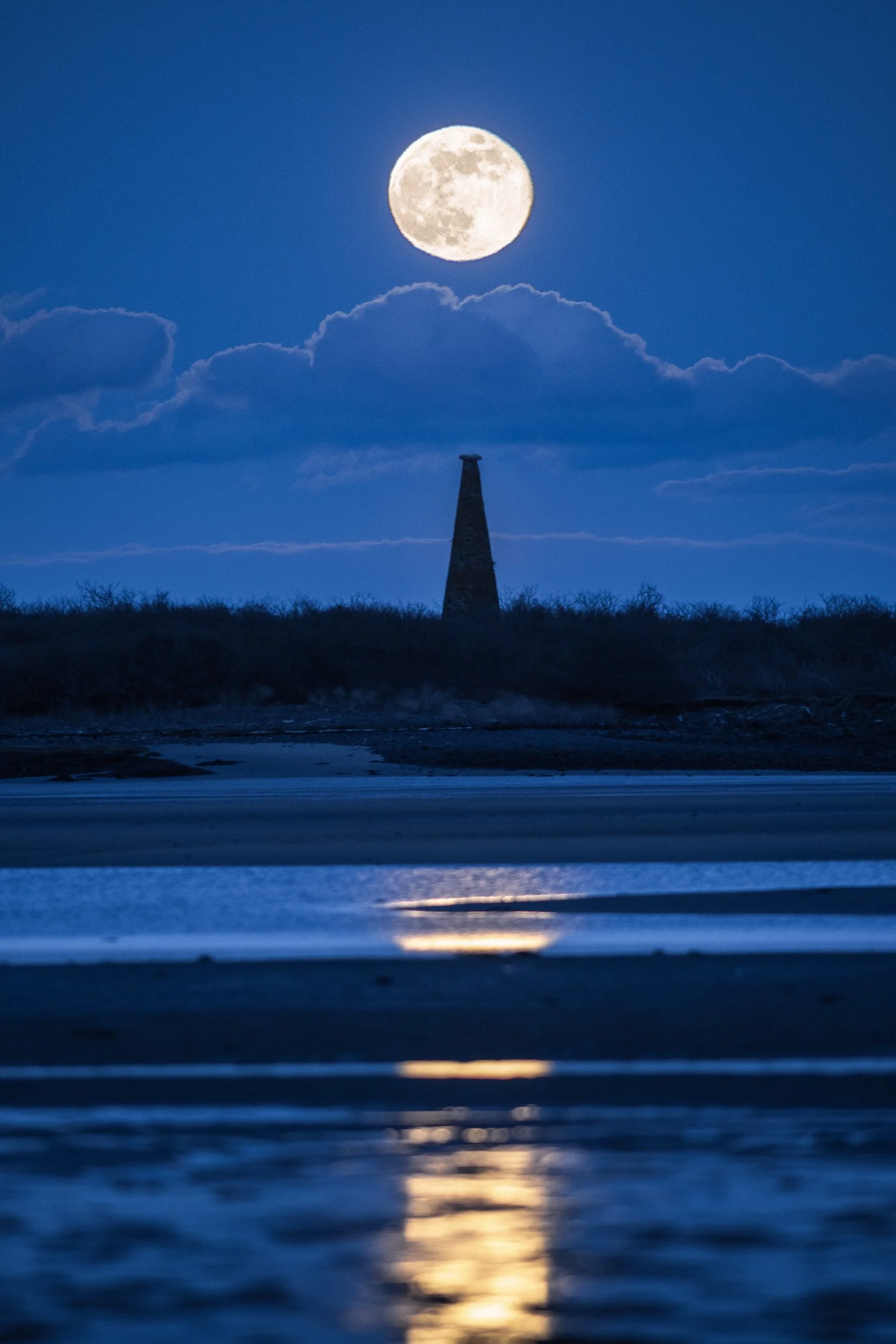 Full moon shining in the night sky with clouds and a silhouette of a chimney on the horizon, and reflections on the water below.