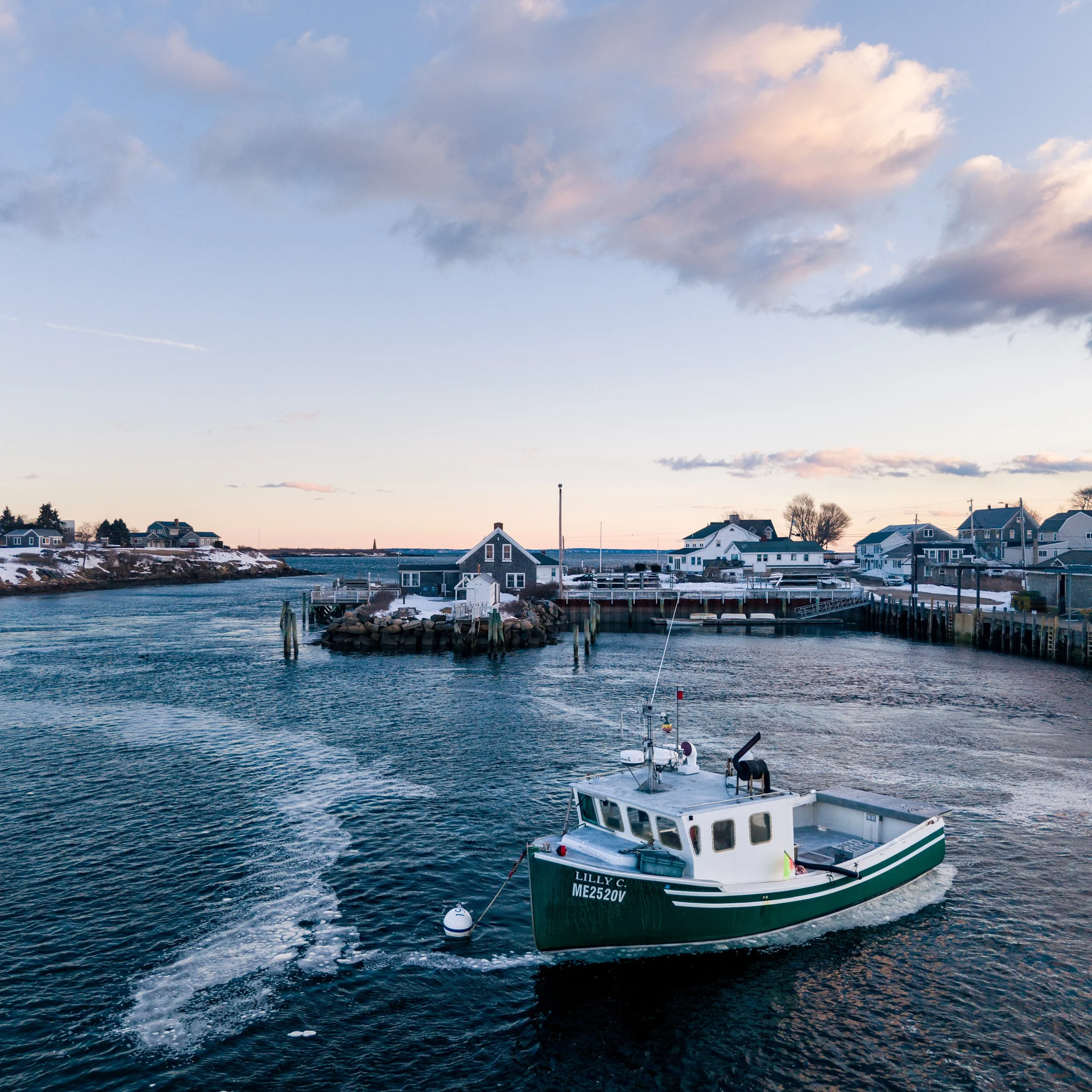 A small boat with the name Lilly C is sailing in a harbor with houses and docks on the shoreline in the background during sunset or sunrise.