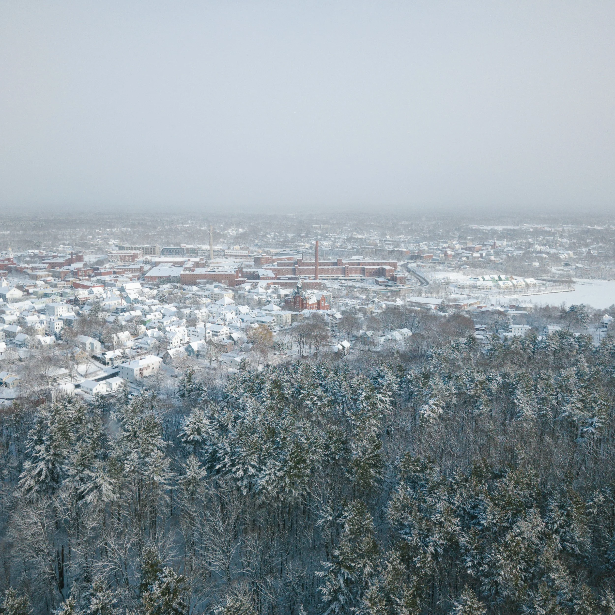 A snow-covered aerial view of Biddeford Maine with many houses, trees, and industrial buildings under a foggy sky.