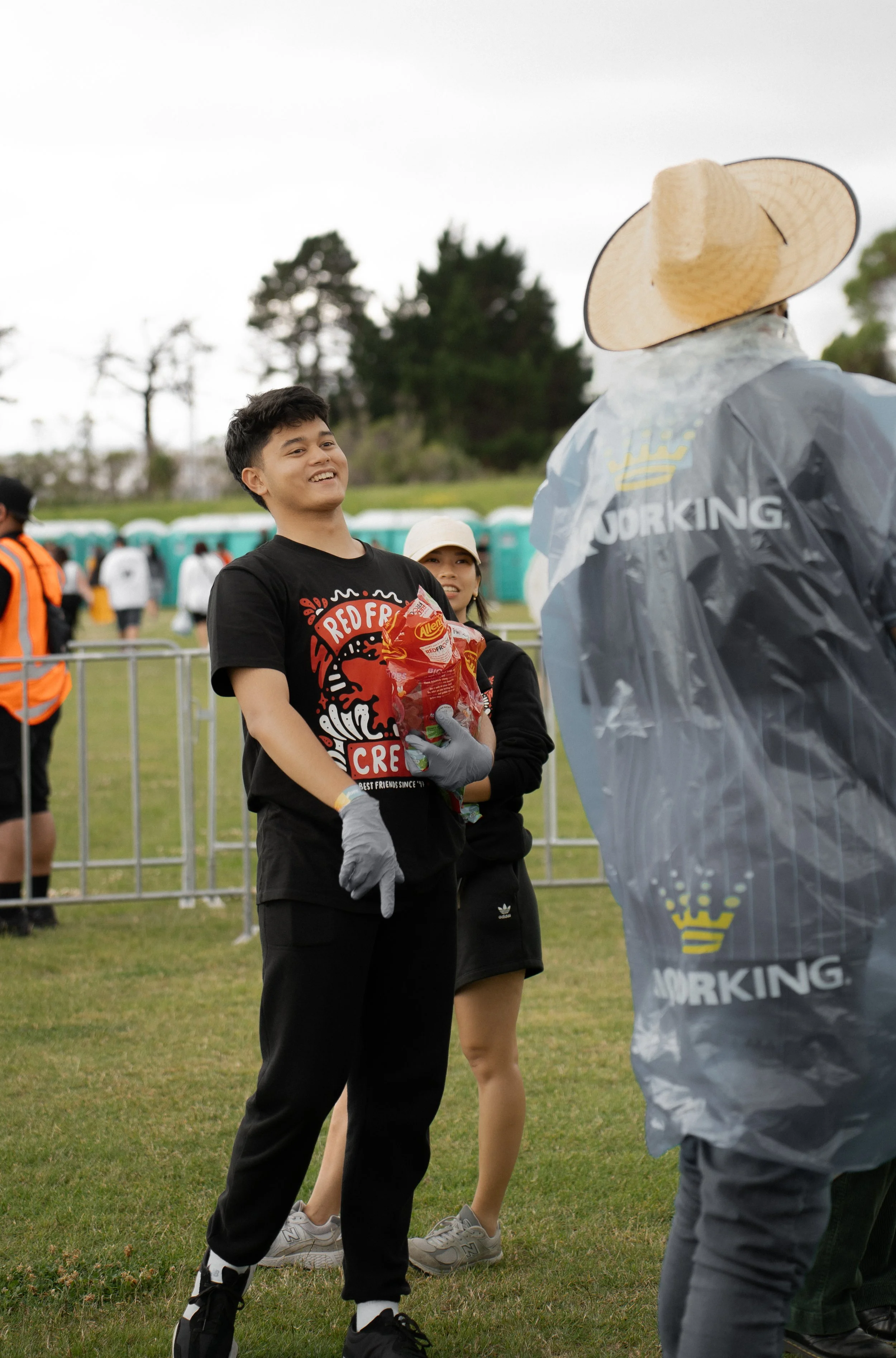 People at an outdoor event, one wearing a black shirt and gloves, holding a snack, another person wearing a plastic rain poncho and straw hat. A line of portable toilets and people are visible in the background.