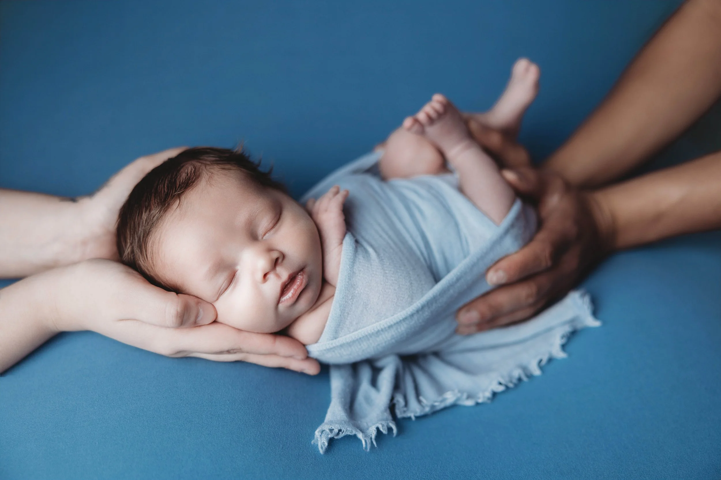 Sleeping newborn baby wrapped in a soft blue swaddle, gently posed in parents’ hands against a solid blue backdrop during a luxury newborn photography session at Helms Photography in Texarkana, Texas.