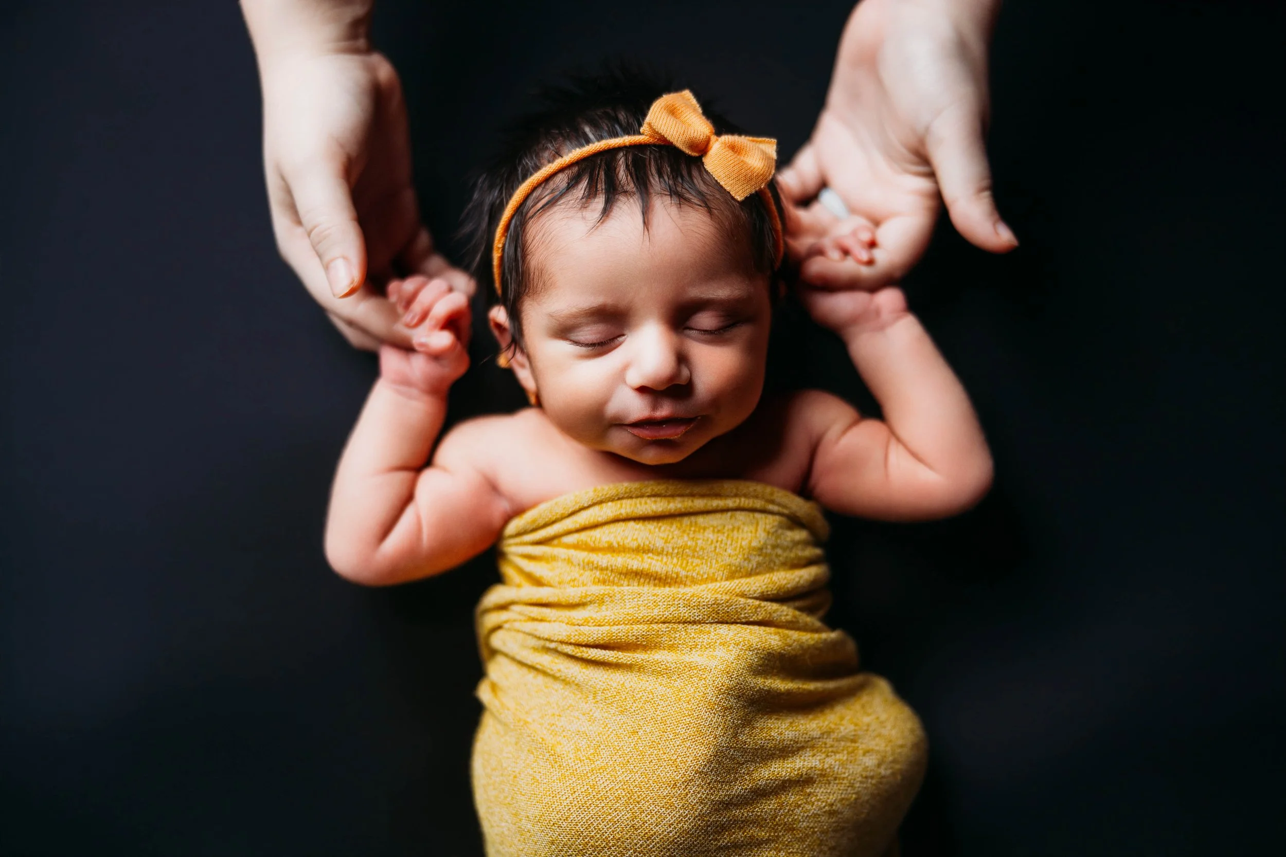 Sleeping newborn baby girl wrapped in a mustard yellow swaddle with a matching bow headband, peacefully posed against a dark backdrop while gently holding her parents’ hands during a luxury newborn session at Helms Photography in Texarkana, Texas.