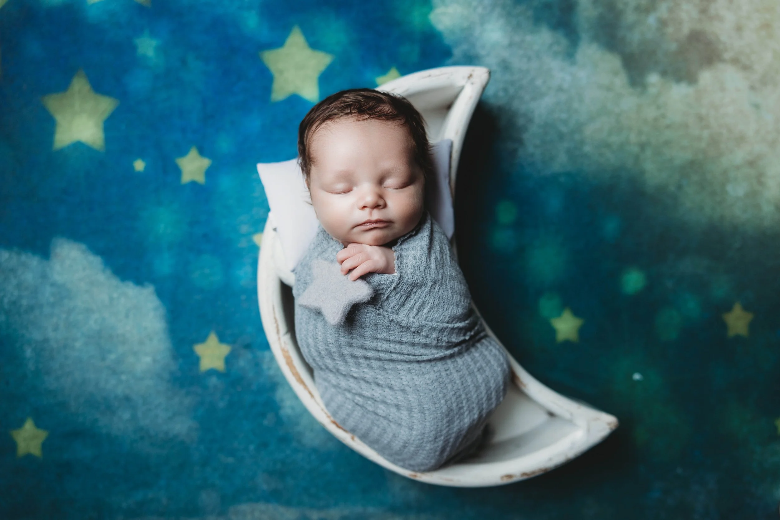 Sleeping newborn baby wrapped in a soft blue swaddle, posed on a white crescent moon prop against a dreamy blue night sky backdrop with clouds and stars, photographed in the luxury newborn studio at Helms Photography in Texarkana, Texas.