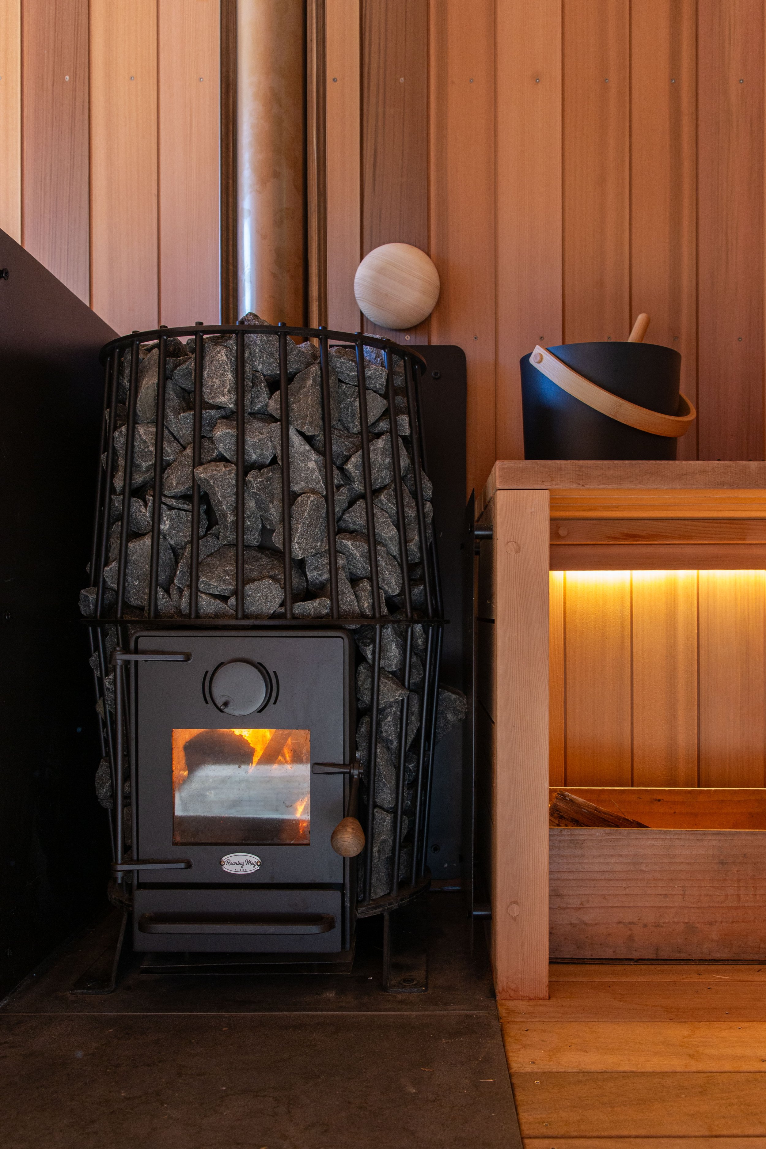 A New Zealand made Roaring Meg wood burning sauna heater burning timber to create the sauna warmth, with a fitted solid cedar bench seat and black water bucket and ladle.