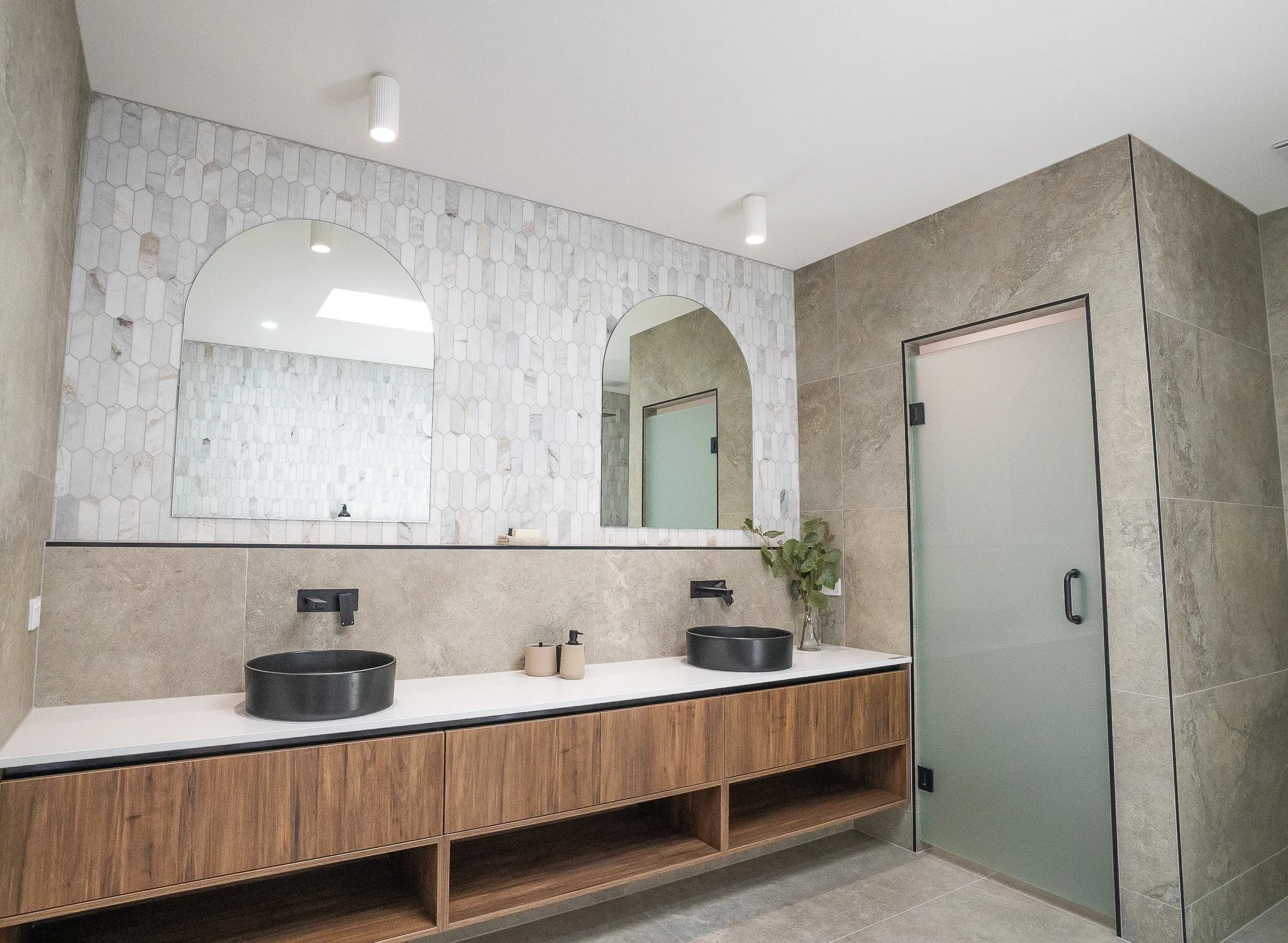 Modern bathroom with double black vessel sinks, large mirrors, wooden vanity, frosted glass door, and decorative tile wall.