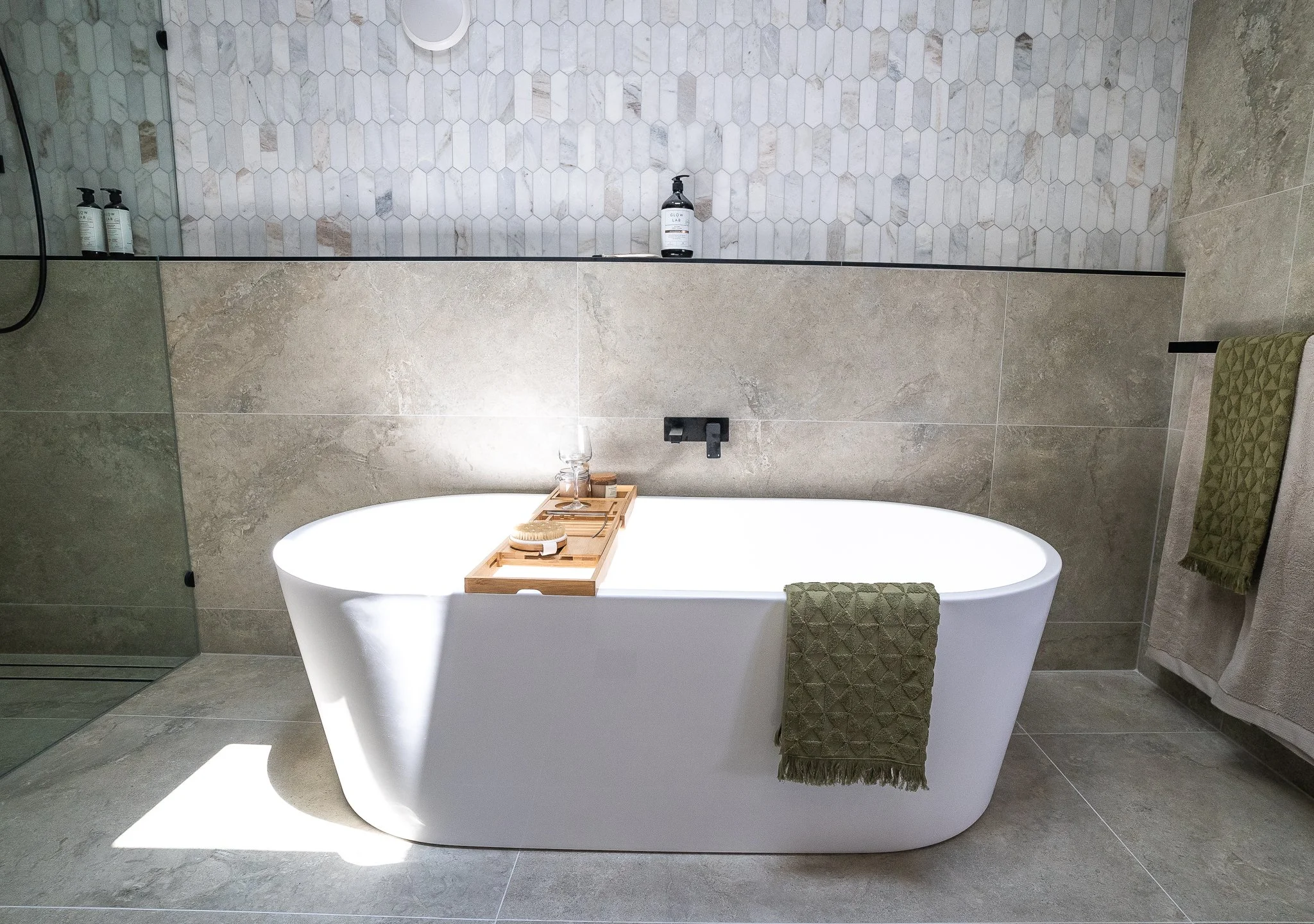 Modern bathroom with a freestanding white bathtub, green towels, gray tiled floor, and a glass shower area with hexagonal tiles in the background.