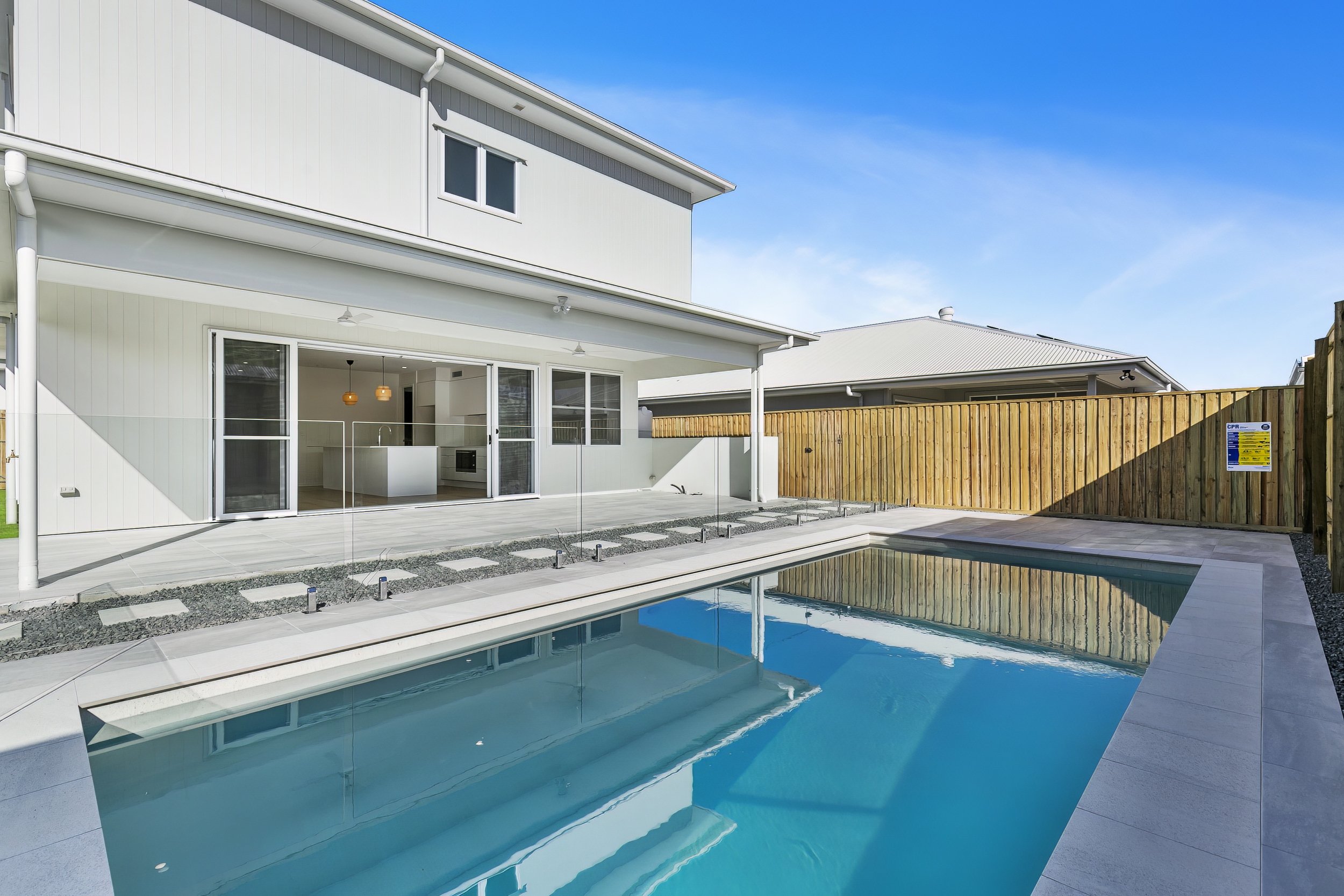 Modern backyard with in-ground swimming pool, patio, and a white two-story house with large sliding glass doors and a wooden fence.