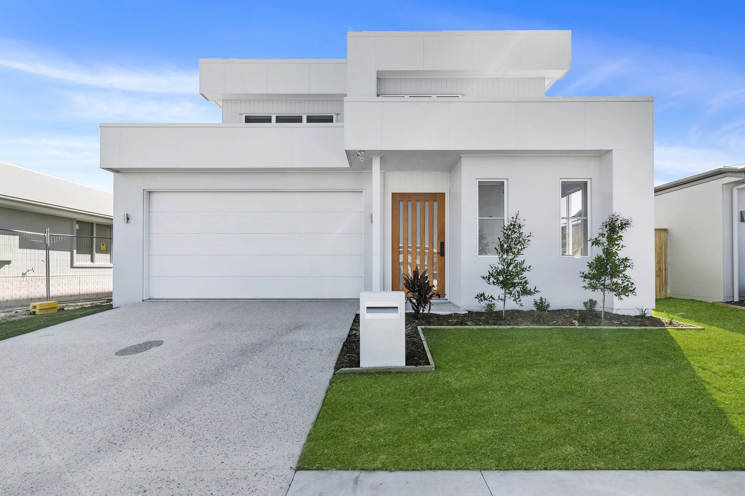 Modern white house with a wooden front door, landscaped yard with grass and small trees, and a concrete driveway, under a blue sky.