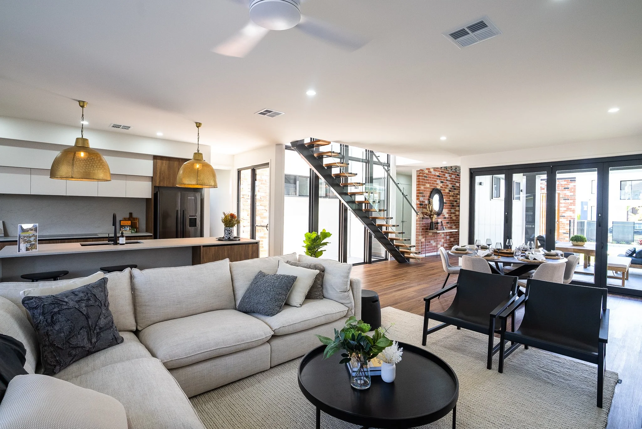 Open-concept living room with a sectional sofa, black chairs, dining table, modern kitchen with gold pendant lights, glass staircase, brick accent wall, and large sliding doors leading outside.