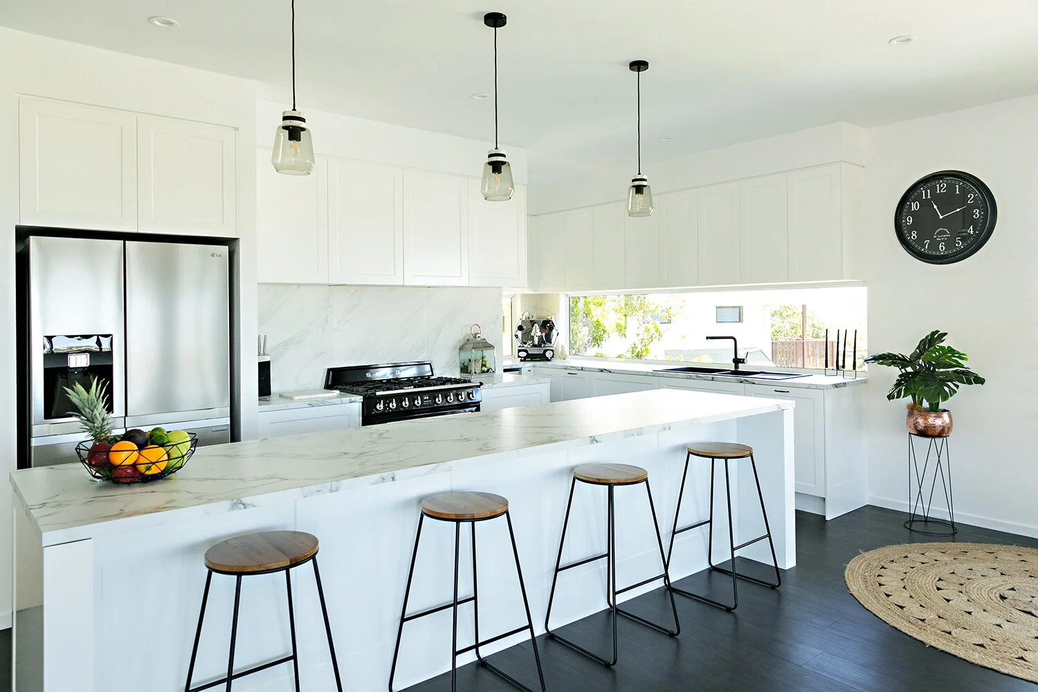 Modern white kitchen with marble island, black and stainless steel appliances, hanging pendant lights, a black clock on the wall, and a potted plant, with a curved jute rug on dark flooring.