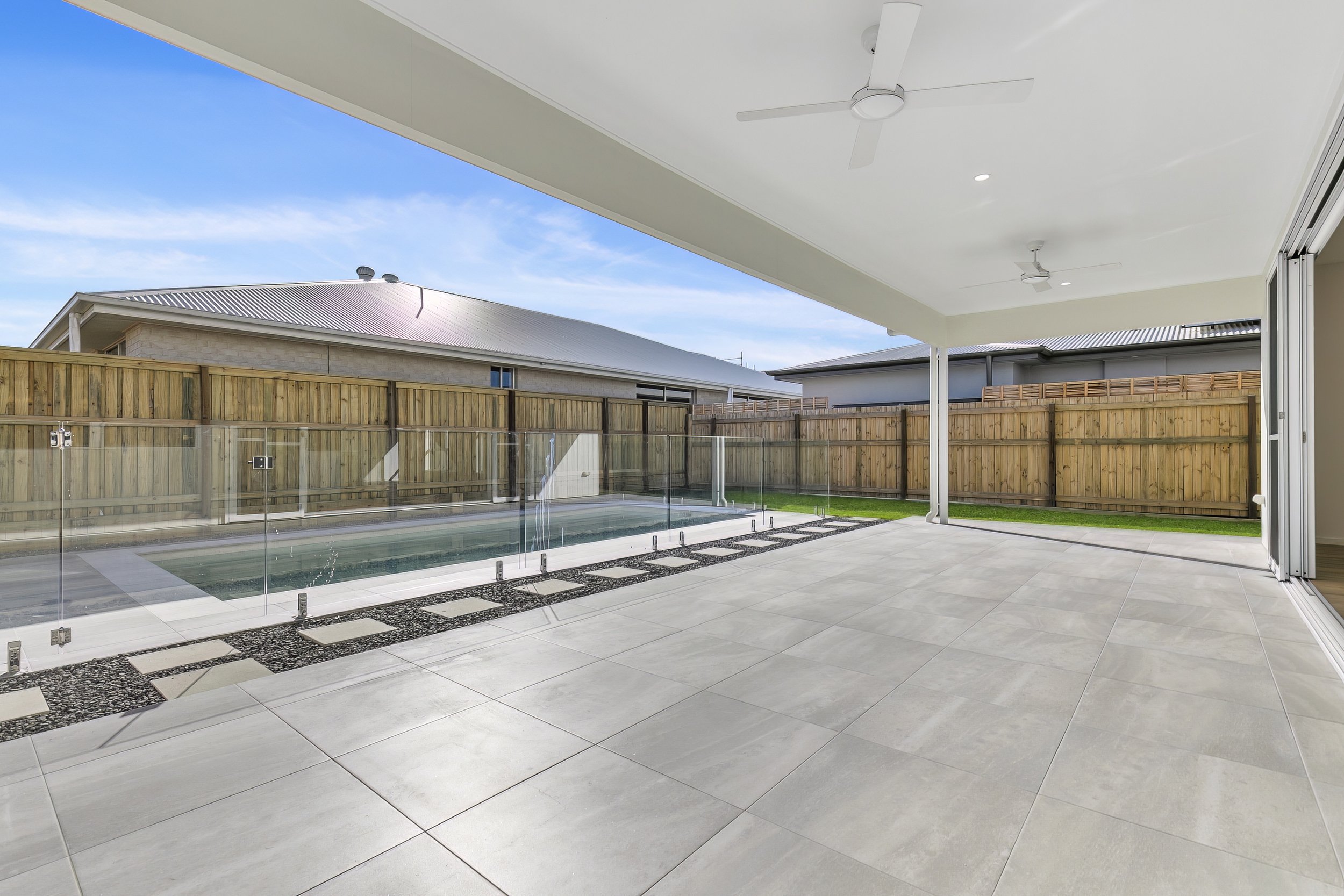 Modern backyard covered patio with large tiled floor, ceiling fans, view of a swimming pool with glass fencing, wooden privacy fence, and neighboring houses under a blue sky.