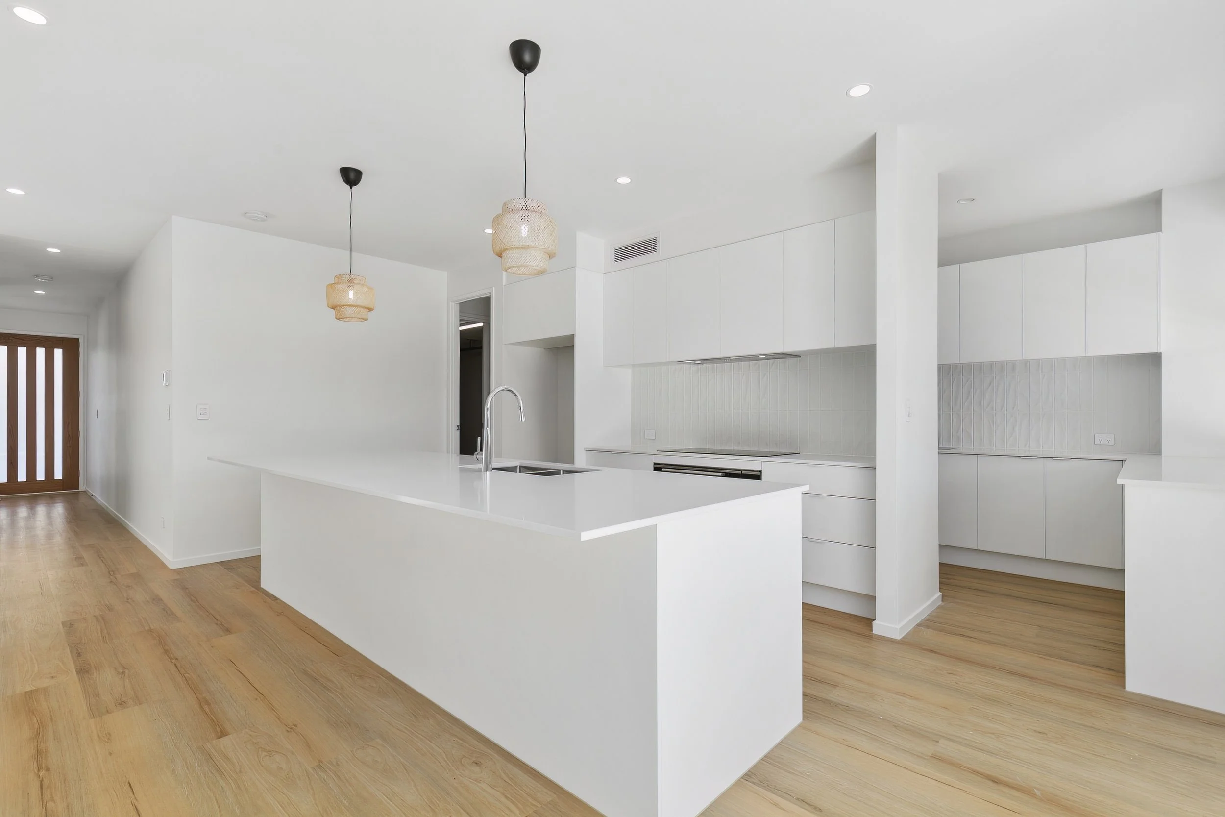 Modern white kitchen with wooden flooring, two hanging pendant lights, and a large island with a sink.
