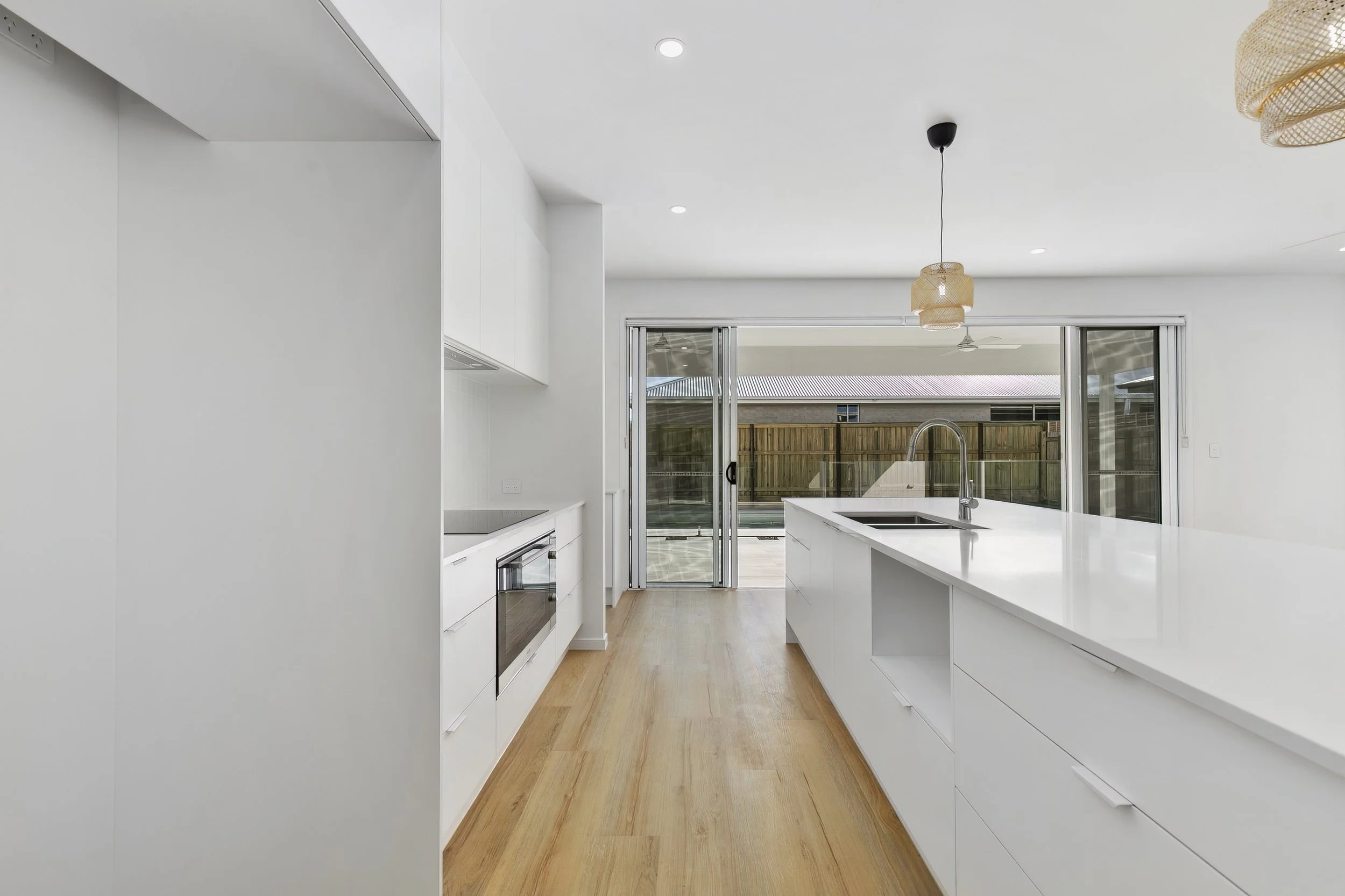Modern white kitchen with island, built-in oven, sliding glass door leading to outdoor patio, and wood flooring.