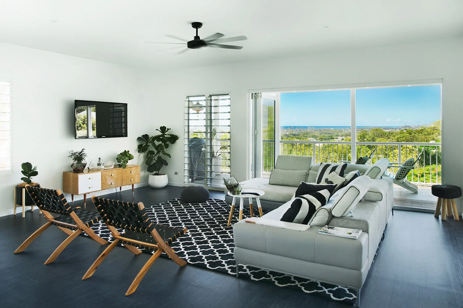 Modern living room with white walls, dark hardwood floor, large sliding glass doors leading to a balcony with outdoor seating, a flat-screen TV mounted on the wall, a wooden sideboard, a white sofa with striped pillows, two black woven chairs, a blac
