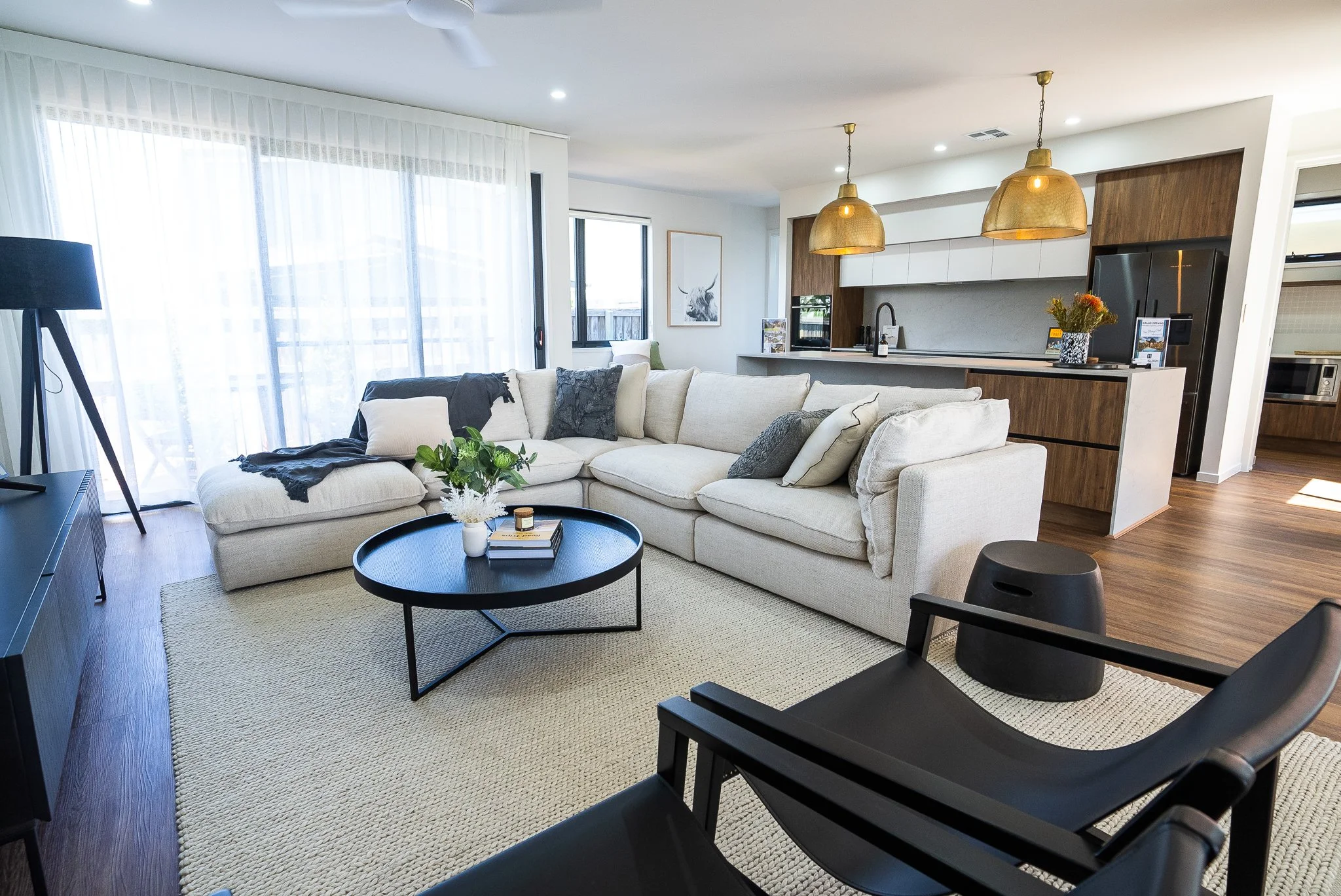 Modern living room with a large white sectional sofa, black coffee table with plants and books, black armchair, area rug, and sliding glass doors with sheer curtains leading outside. Open kitchen with wooden cabinets and gold pendant lights.