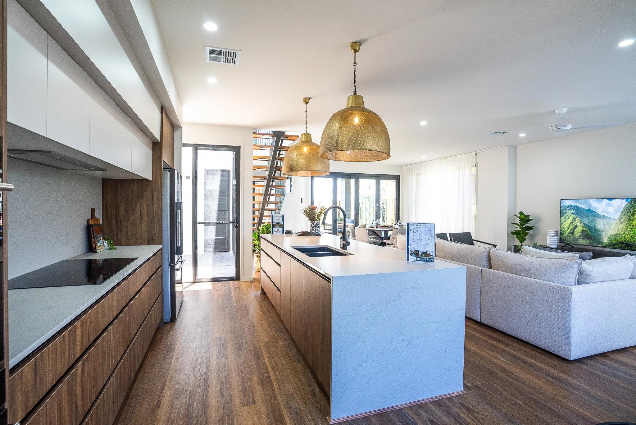 Modern open-concept kitchen and living room with wooden flooring, white walls, a central island with a sink, beige sectional sofa, flat-screen TV, dining table near large windows, and gold pendant lights.