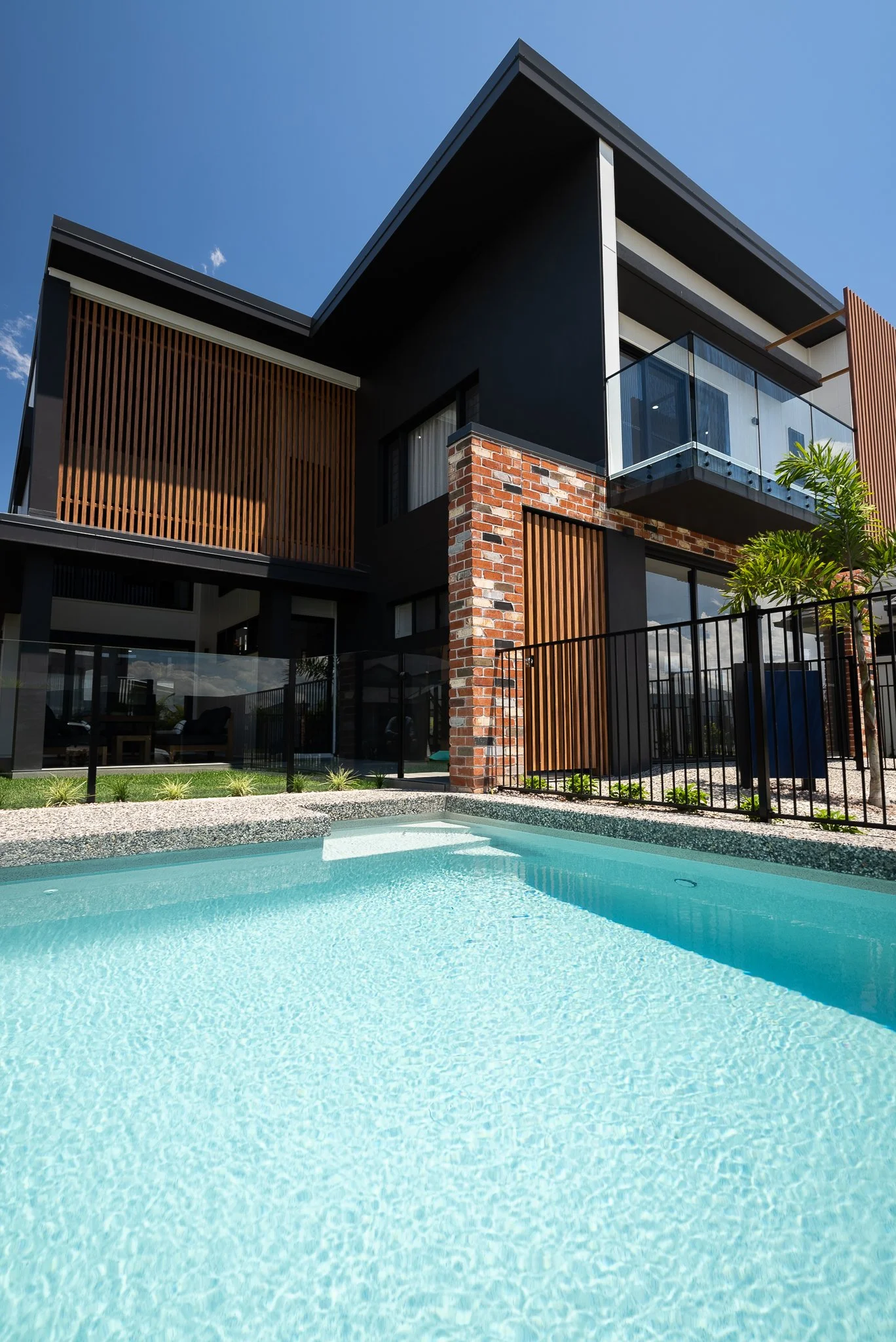 Modern multi-story house with a swimming pool in the backyard, featuring brick and wood accents, glass balcony, and clear blue sky.