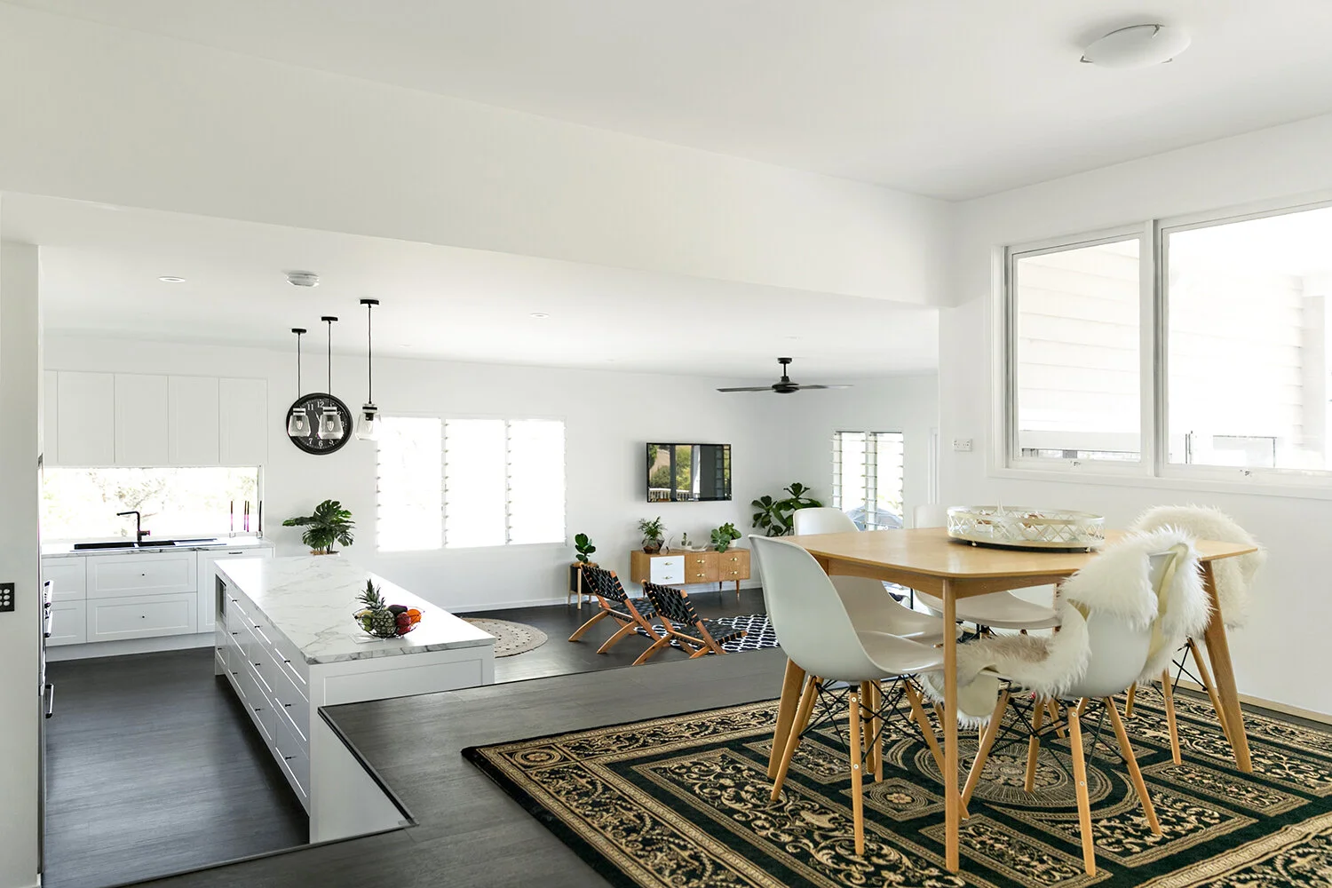 Open-concept modern kitchen and dining area with white walls, wooden dining table with white and fur-covered chairs, and dark hardwood floors.
