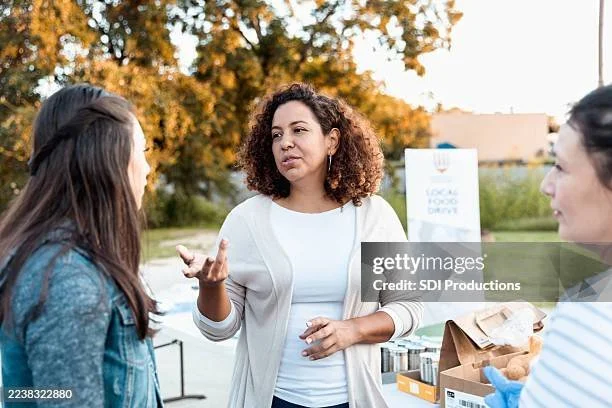 Three women having a conversation outdoors, with a food drive sign and tables with food items in the background, during late afternoon.
