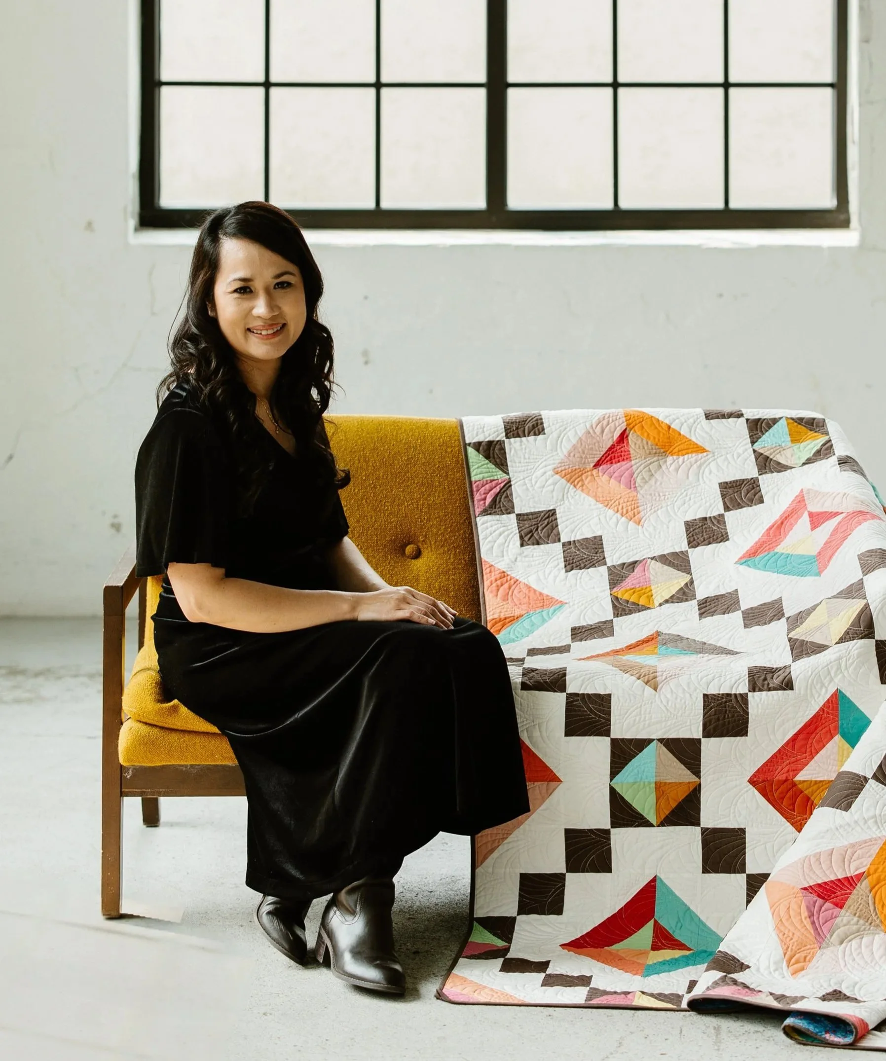 A woman with dark hair, wearing a black dress and white shoes, sitting on a yellow chair near a window with black framing. A colorful quilt with geometric patterns is draped over a piece of furniture beside her.