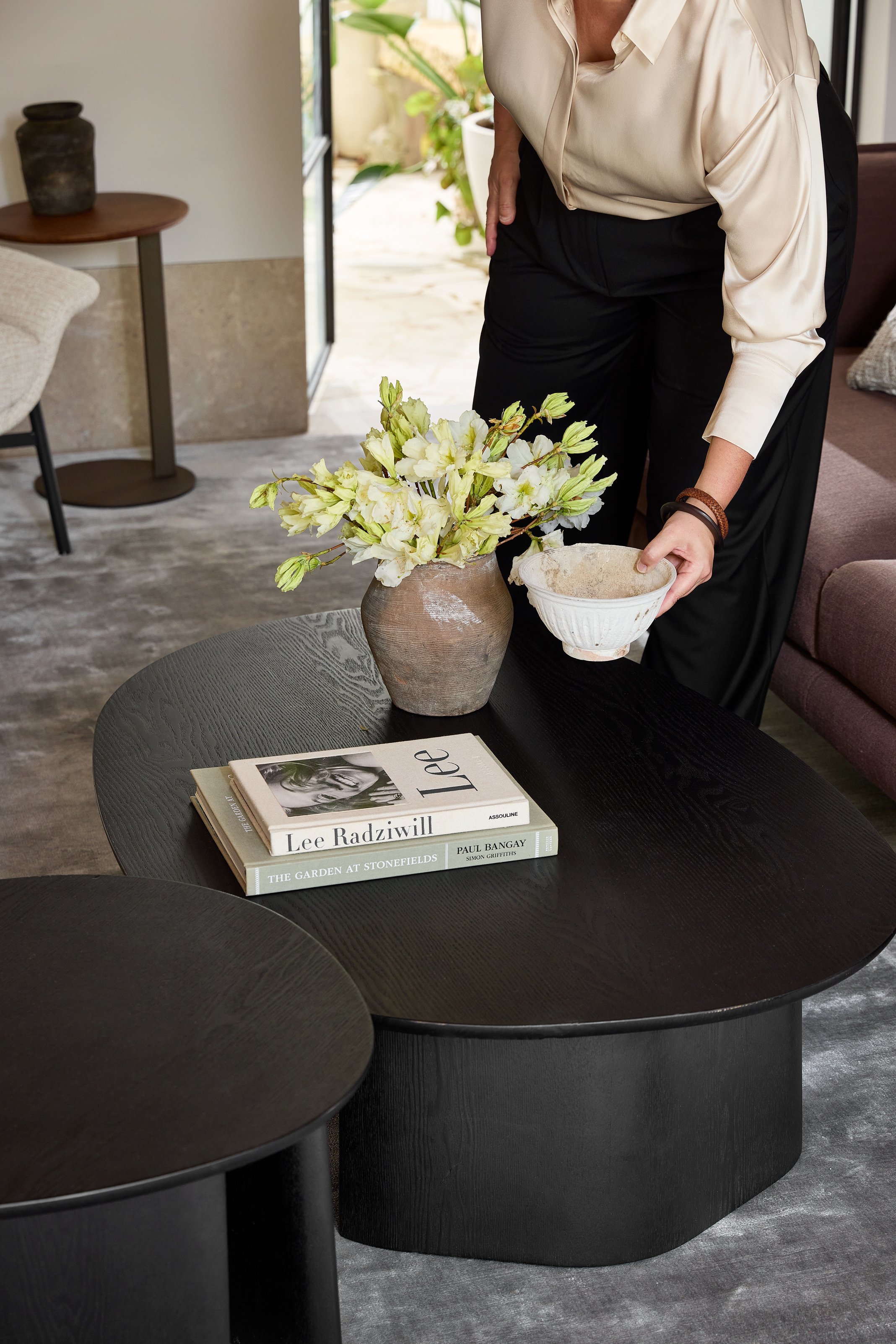 A woman arranging a flower vase with white flowers on a black coffee table, with books titled 'Lee Radziwill' and 'The Garden at Stonefields' nearby, in a modern living room with a gray rug and furniture.