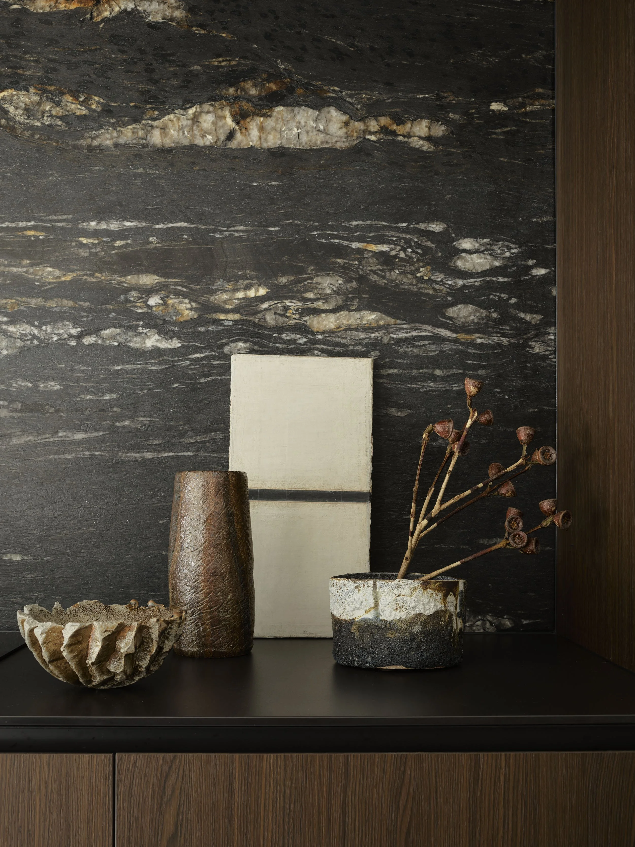 Decorative arrangement of vases and objects on a dark surface against a textured dark stone wall. Includes a round stone bowl, a tall cylindrical brown textured vase, a rectangular beige sculpture, and a small round pot with dried plant stems.