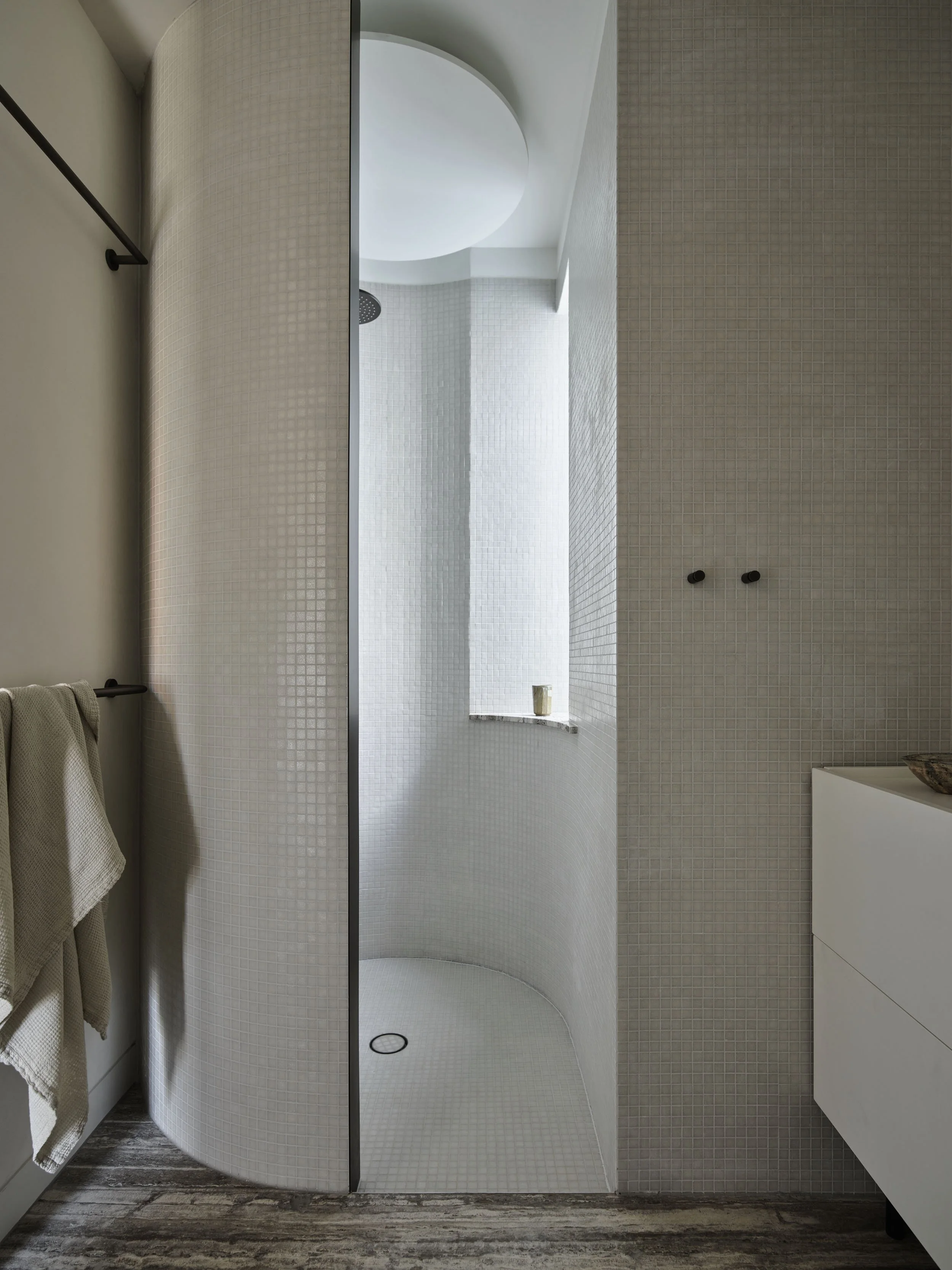 Minimalist, curved walk-in shower with white mosaic tiles, a built-in shelf with a small yellow cup, and natural light from a narrow window.