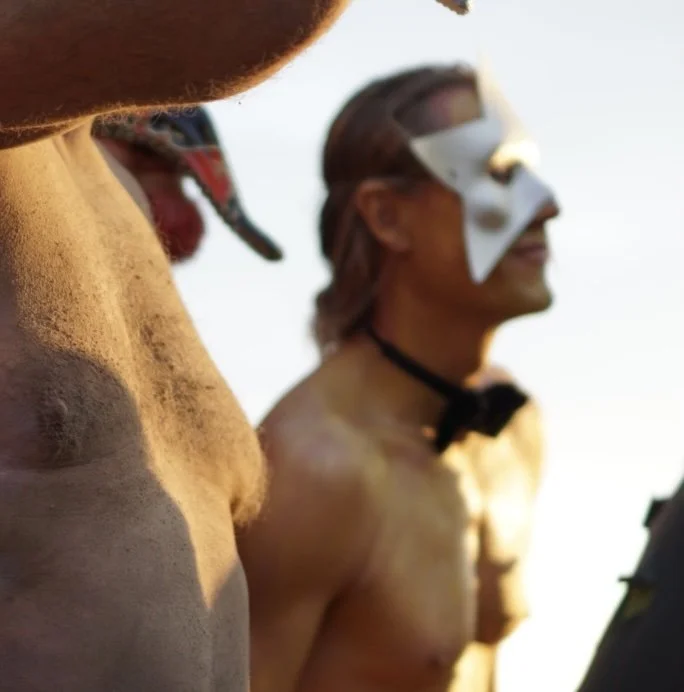 To mennesker uden overtøj, en i en masker og en uden, med fokus på deres overkroppe i solnedgangen.