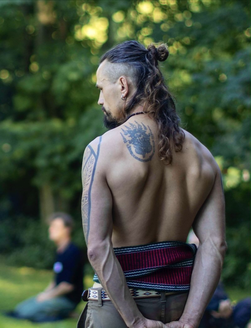En shirtløs mand med tatoveringer på ryggen og armen, med langt, bølget hår og skæg, set bagfra i en grøn udendørs setting.