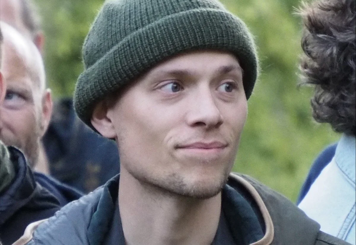 En ung mand har på en grøn hue og en sort jakke, og der står en gruppe mennesker omkring ham udendørs.