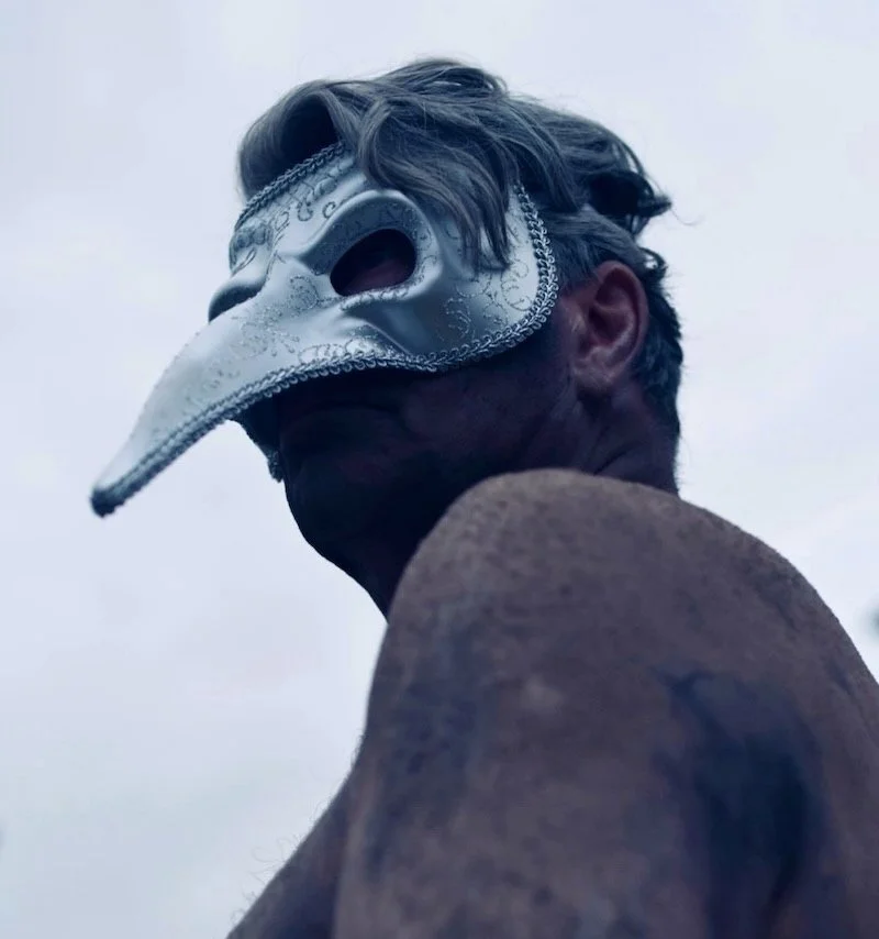 Person iført en hvid maske, der ligner en plague doctor-maskefasade, set fra siden under en overskyet himmel.