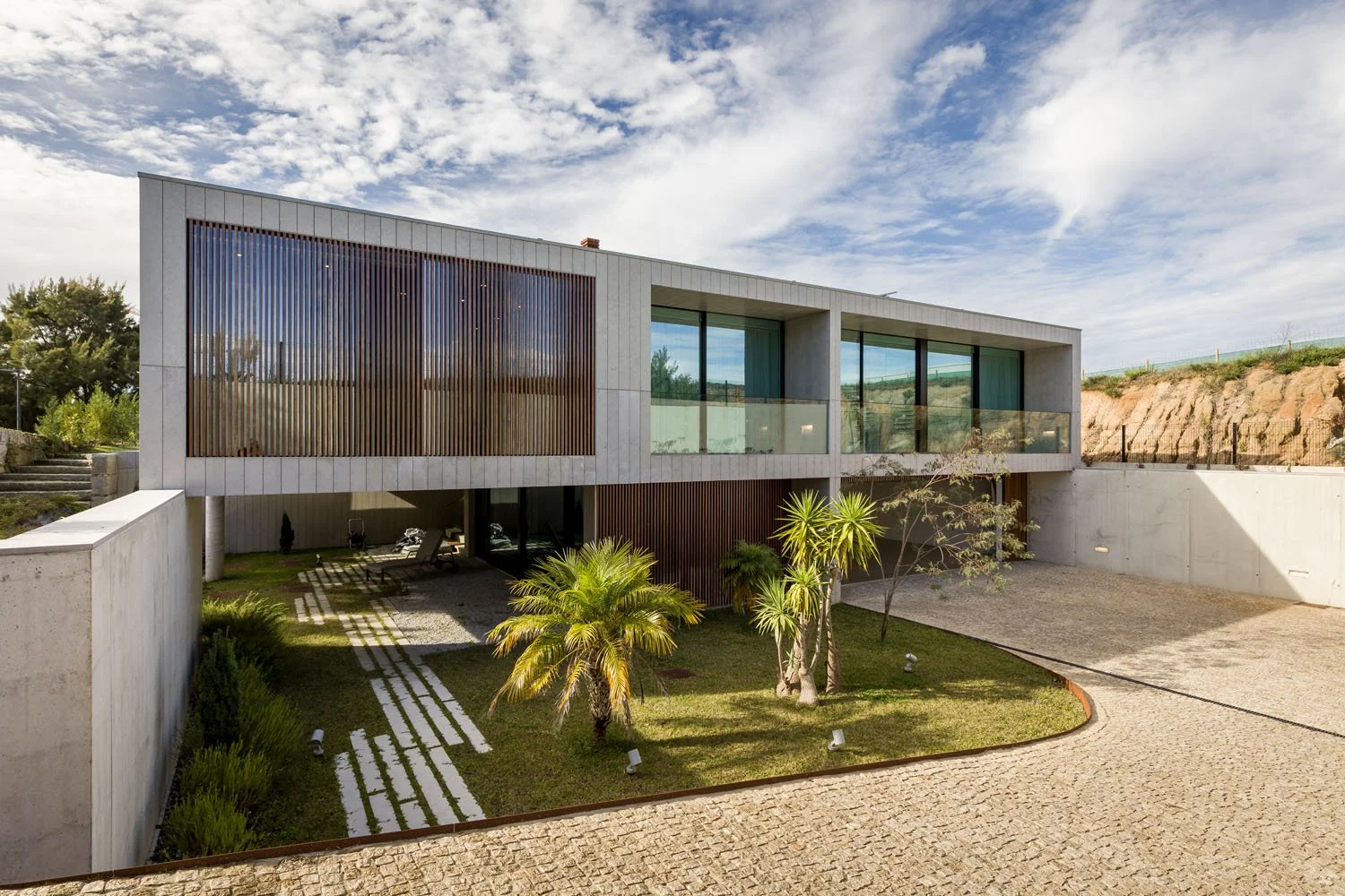 Residência moderna com fachada de vidro e elementos de madeira, jardim com árvores e passeio de pedra.
