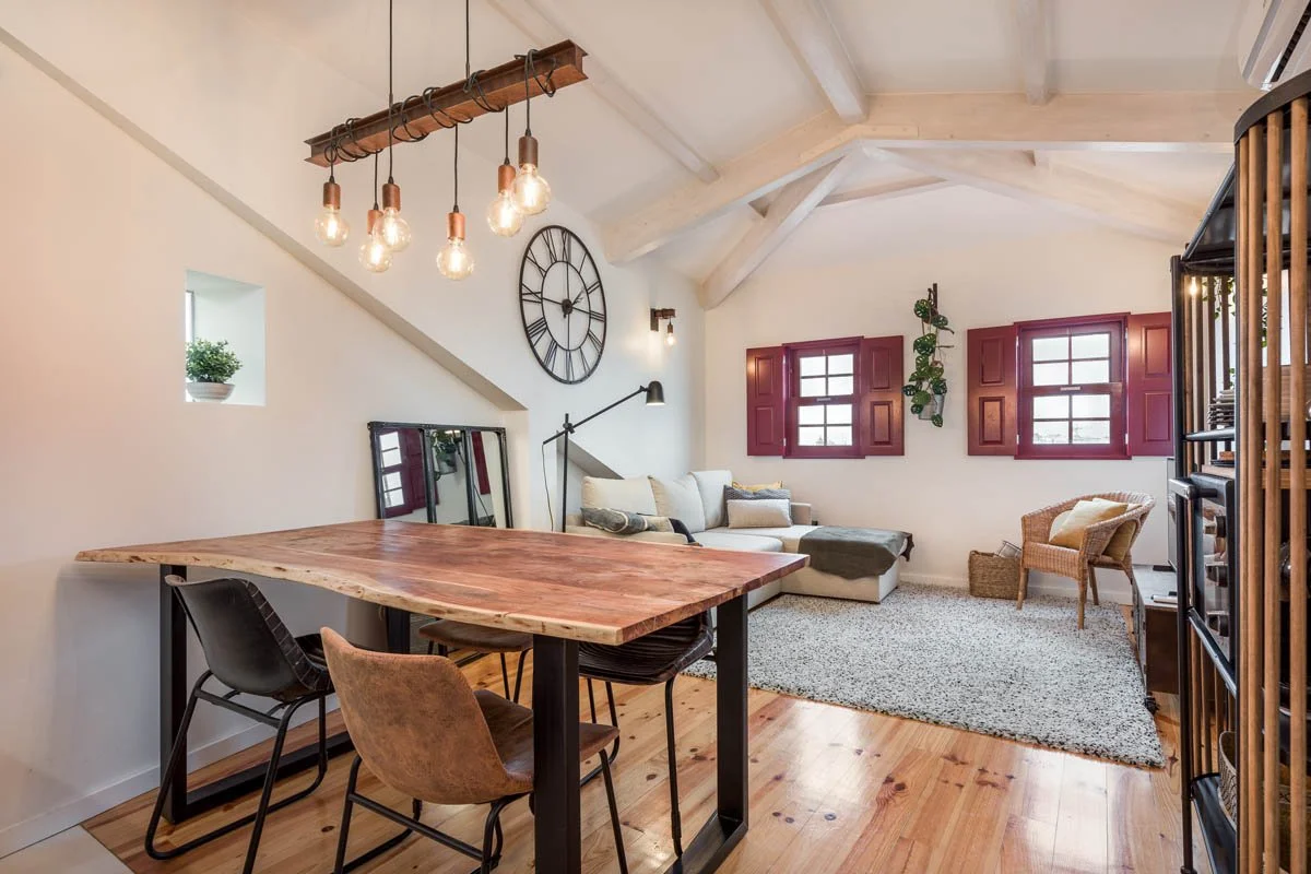 Interior de uma sala de estar com janela, sofá cinza, mesa de jantar de madeira, iluminação pendente com lâmpadas de filamento, e detalhes decorativos em estilo rústico.