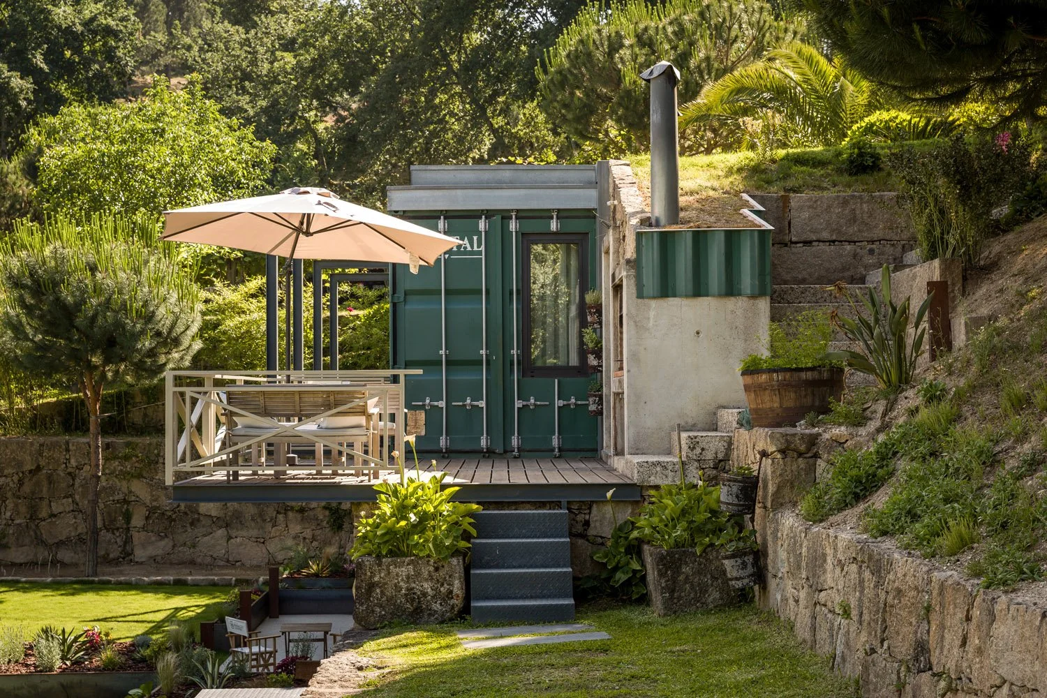 Edícula de estrutura de containers de metal verde com área de lazer com mesa, cadeiras e guarda-sol, rodeada de jardim e vegetação