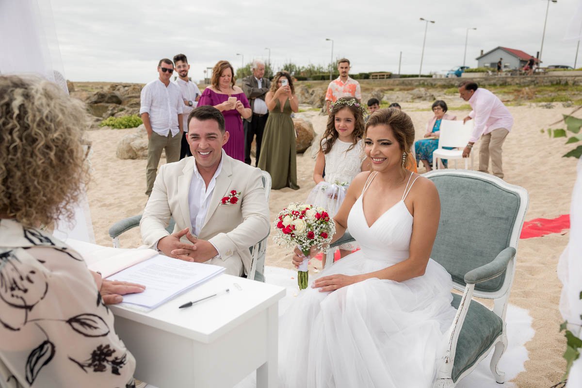 Casal de noivos sorrindo durante a cerimônia de casamento na praia, com convidados ao fundo.