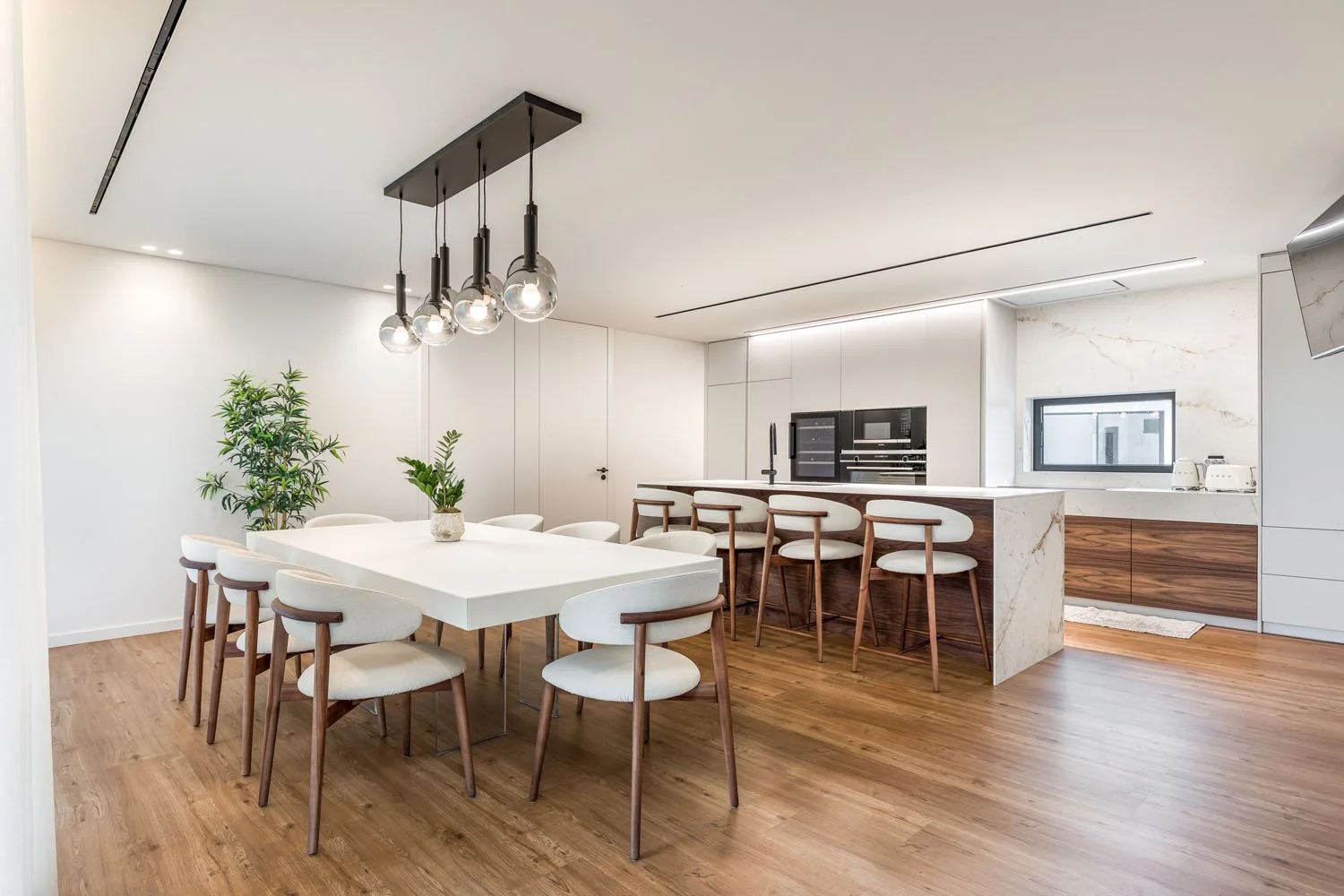 Cozinha moderna com bancada de mármore branco e banquetas de madeira, mesa de jantar branca com cadeiras de tecido claro, piso de madeira, iluminação pendente de vidro, plantas verdes e eletrodomésticos embutidos.